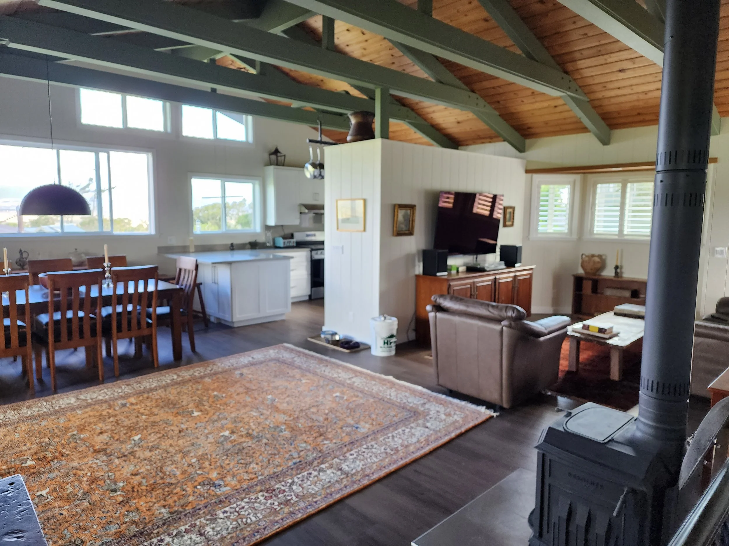 Interior of a living room with a wooden ceiling with exposed beams, a large patterned area rug, a flat-screen TV on a wooden stand, a brown leather armchair, a wooden coffee table, and a wood stove in the foreground. There is a dining area with a woo