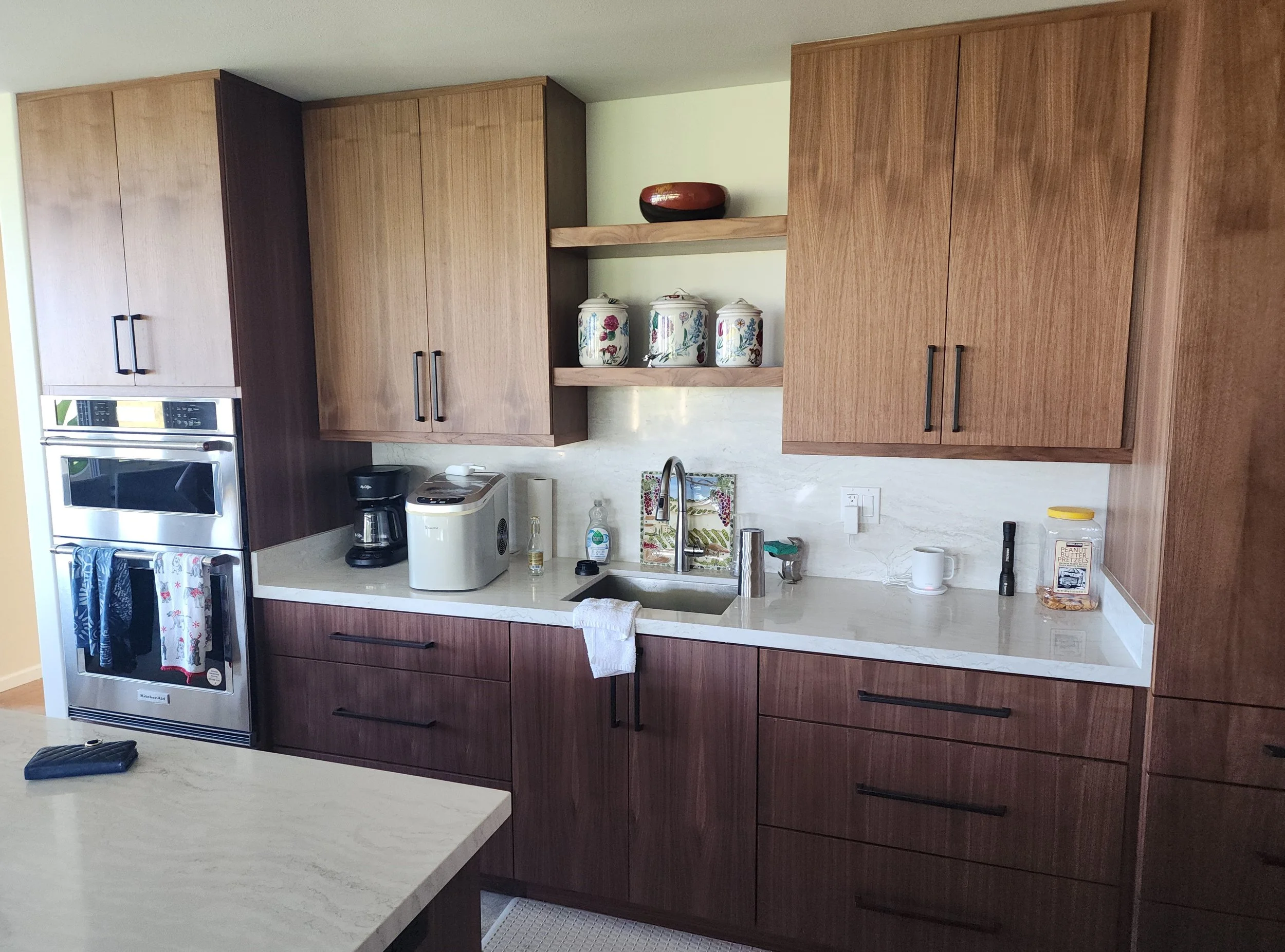 Kitchen with wooden cabinets, a white countertop, a sink, and various kitchen items including a coffee maker, rice cooker, and jars on open shelves.