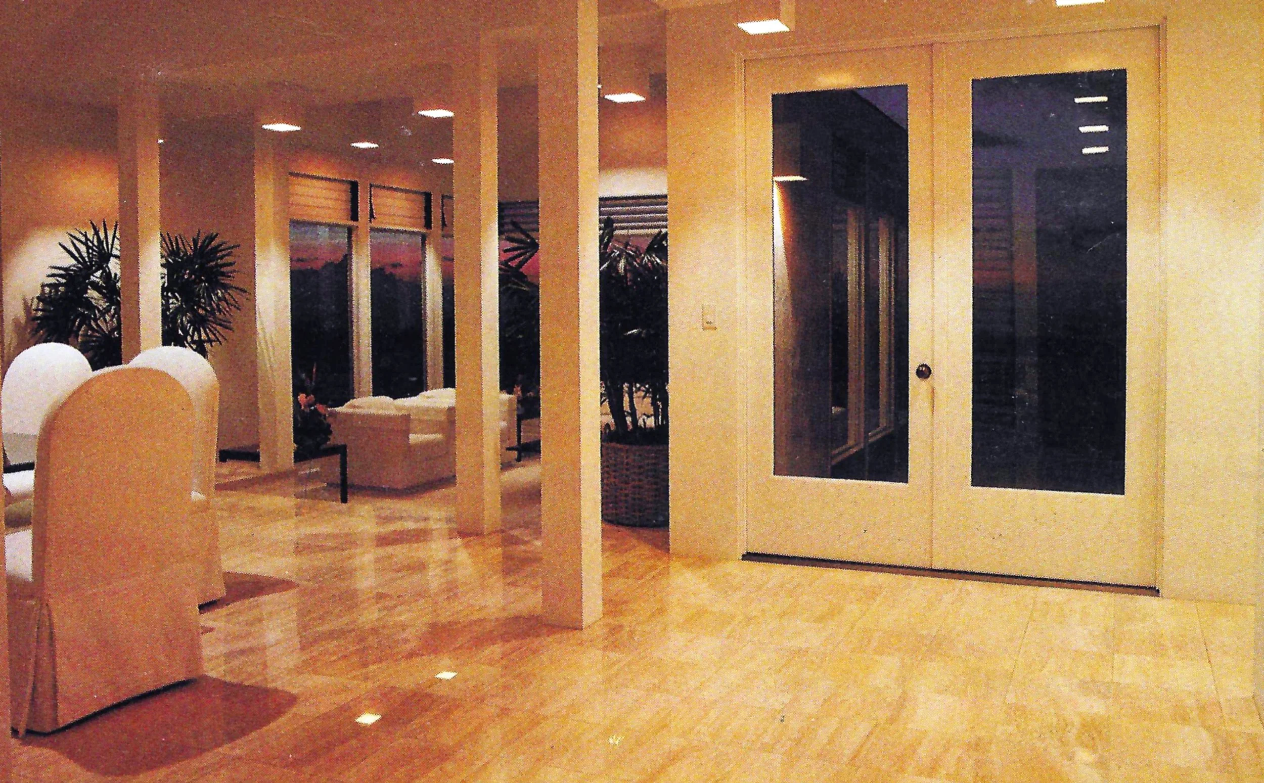 A spacious, well-lit interior room with light-colored wood floors and walls. There are large windows with views of a sunset or dusk sky outside, and indoor plants are visible. The room contains a white armchair, a white sofa, and decorative moon-shap