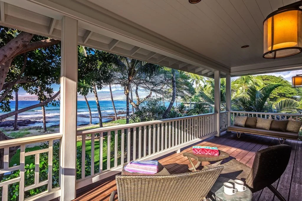 A covered porch overlooking a tropical beach with palm trees, ocean waves, and blue sky, decorated with outdoor furniture and warm lighting.