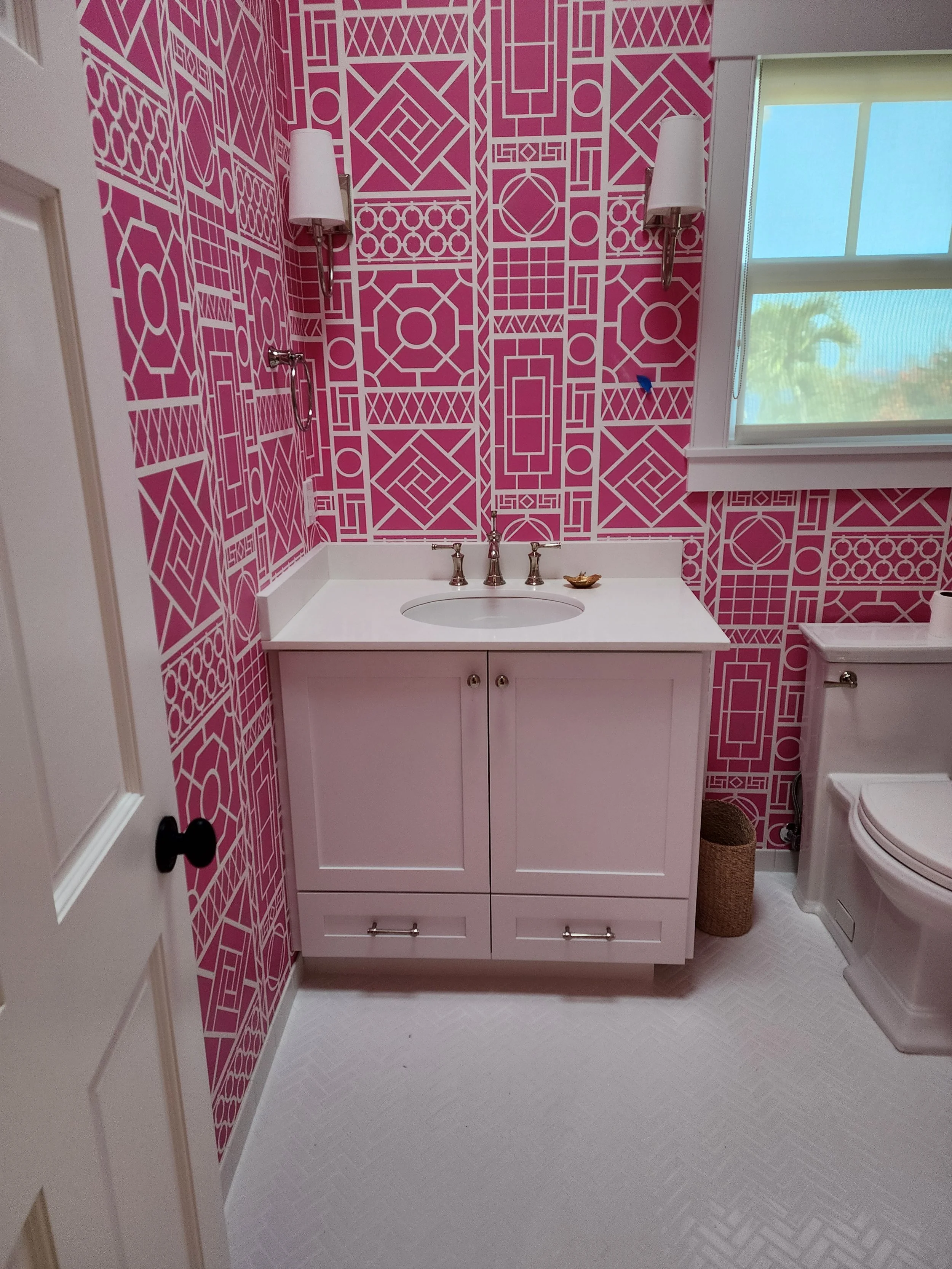 A bathroom with pink patterned wallpaper, a white vanity with a sink, silver faucet, and cabinet, paired with wall-mounted white light fixtures on either side, a window showing outdoor greenery, a toilet, a brown basket, and a white textured floor.