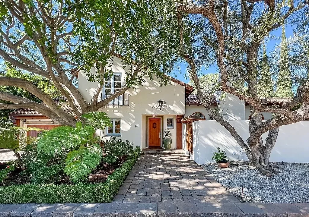 Front view of a white house with a brick pathway leading to the front door, surrounded by trees and garden plants.