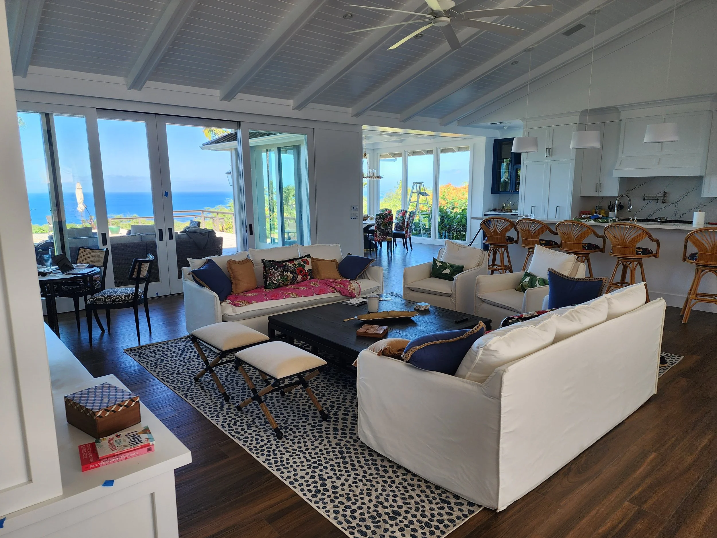 A bright living room with white sofas, colorful pillows, a black coffee table, and large glass sliding doors opening to a patio with ocean views.