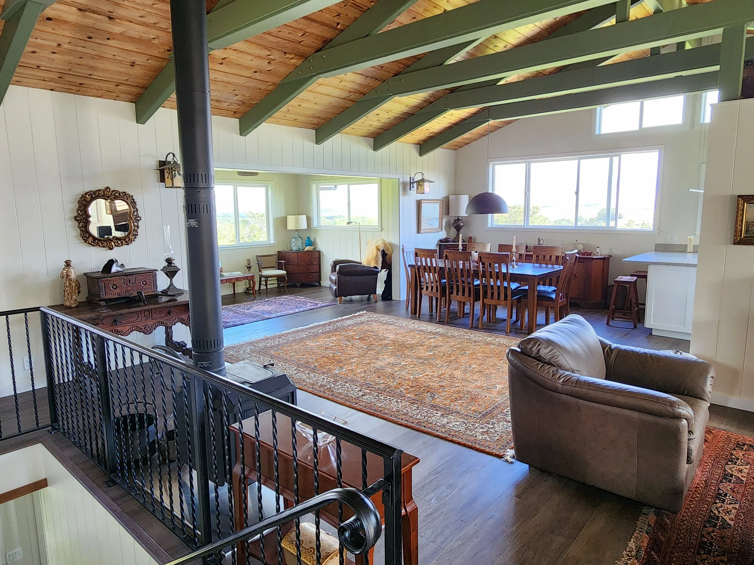 Living room with wooden ceiling beams, large windows, cream walls, hardwood floors, a patterned rug, leather armchair, wooden dining table with chairs, vintage sideboard, and decorative items.