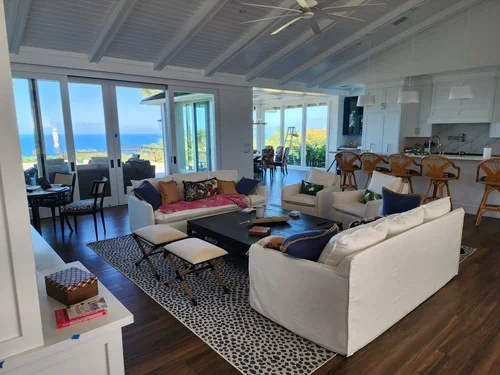 Open-concept Big Island home interior with white sofas, vaulted ceiling, kitchen seating, dark wood floors, and ocean views through large windows.