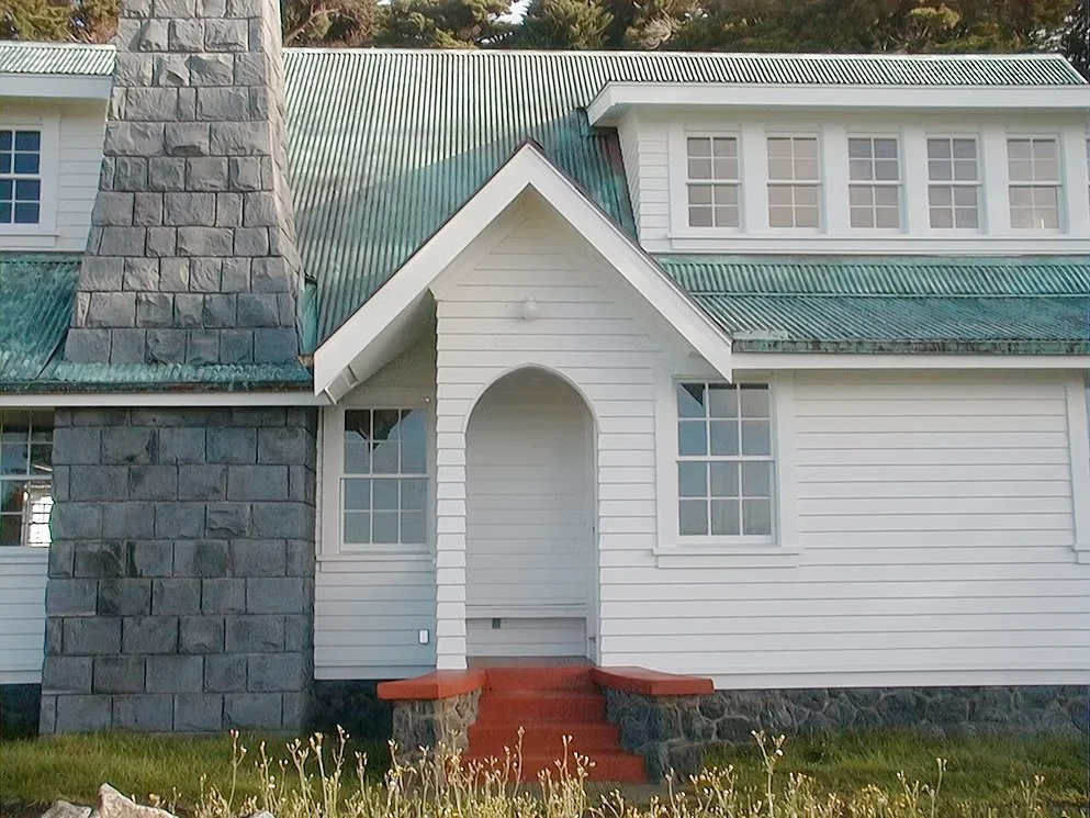 Front view of a house with white siding, a green metal roof, and a large stone chimney.