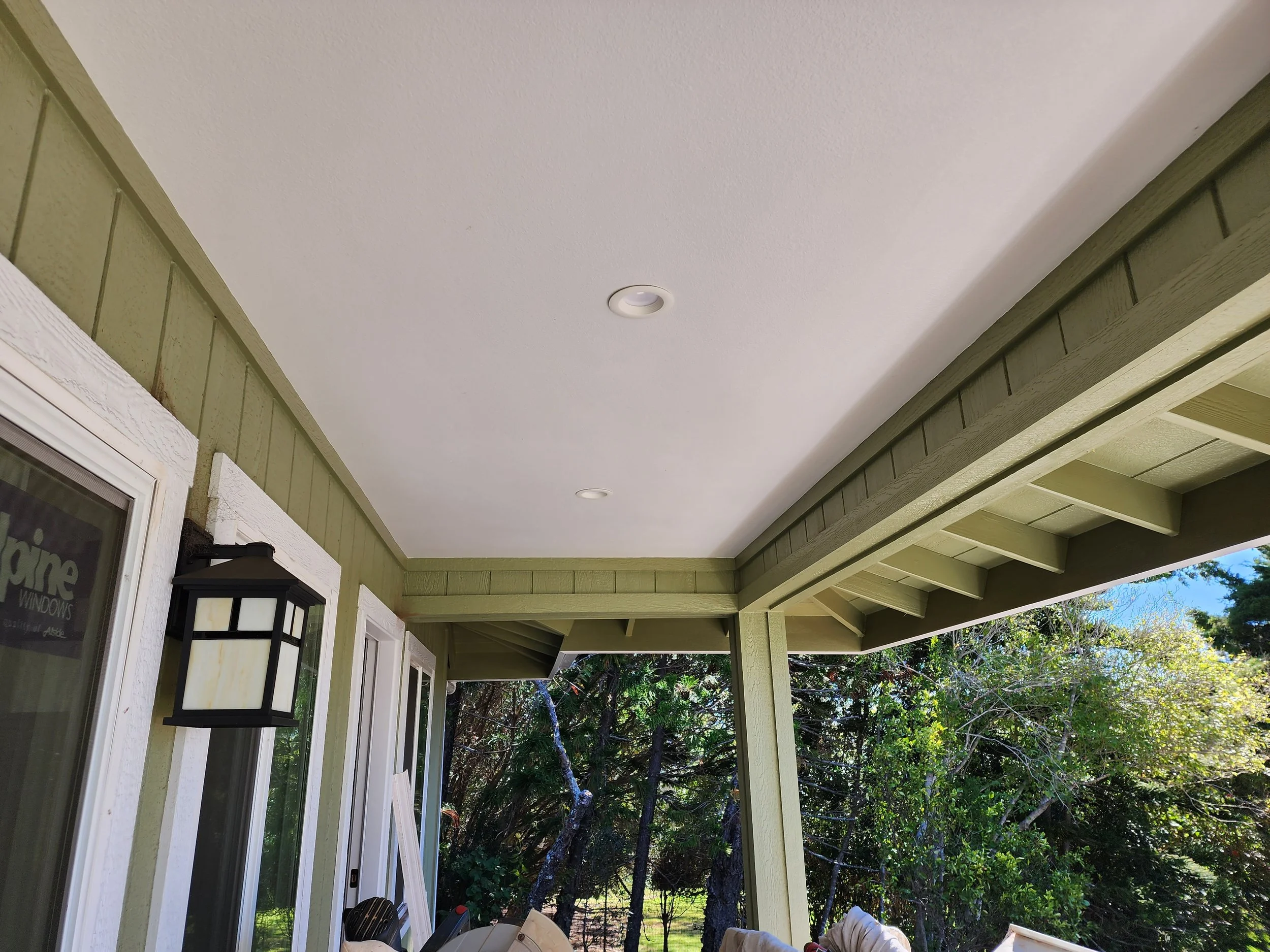 Covered porch with green siding, a black outdoor wall lantern, sliding glass door, and trees in the background.