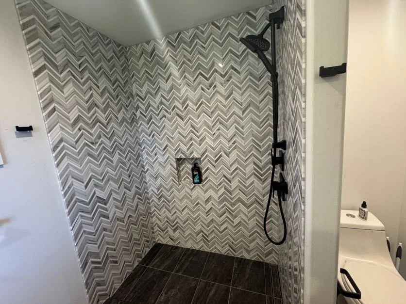 Modern walk-in shower with chevron-patterned tile walls, black handheld showerhead, and built-in shampoo niche. Part of a bathroom with a toilet nearby.