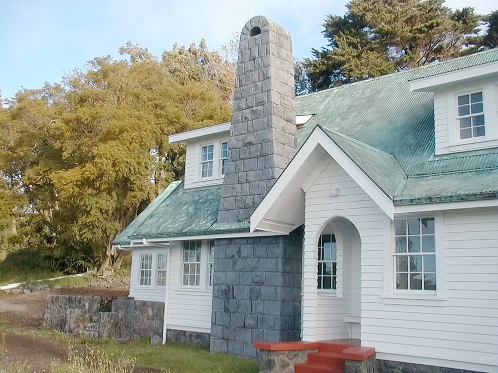 White house with green roof, grey stone chimney, and front steps, surrounded by trees.