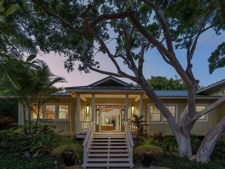Big Island custom home exterior with covered entry, metal roof, mature tropical landscaping, and a large shade tree at dusk.