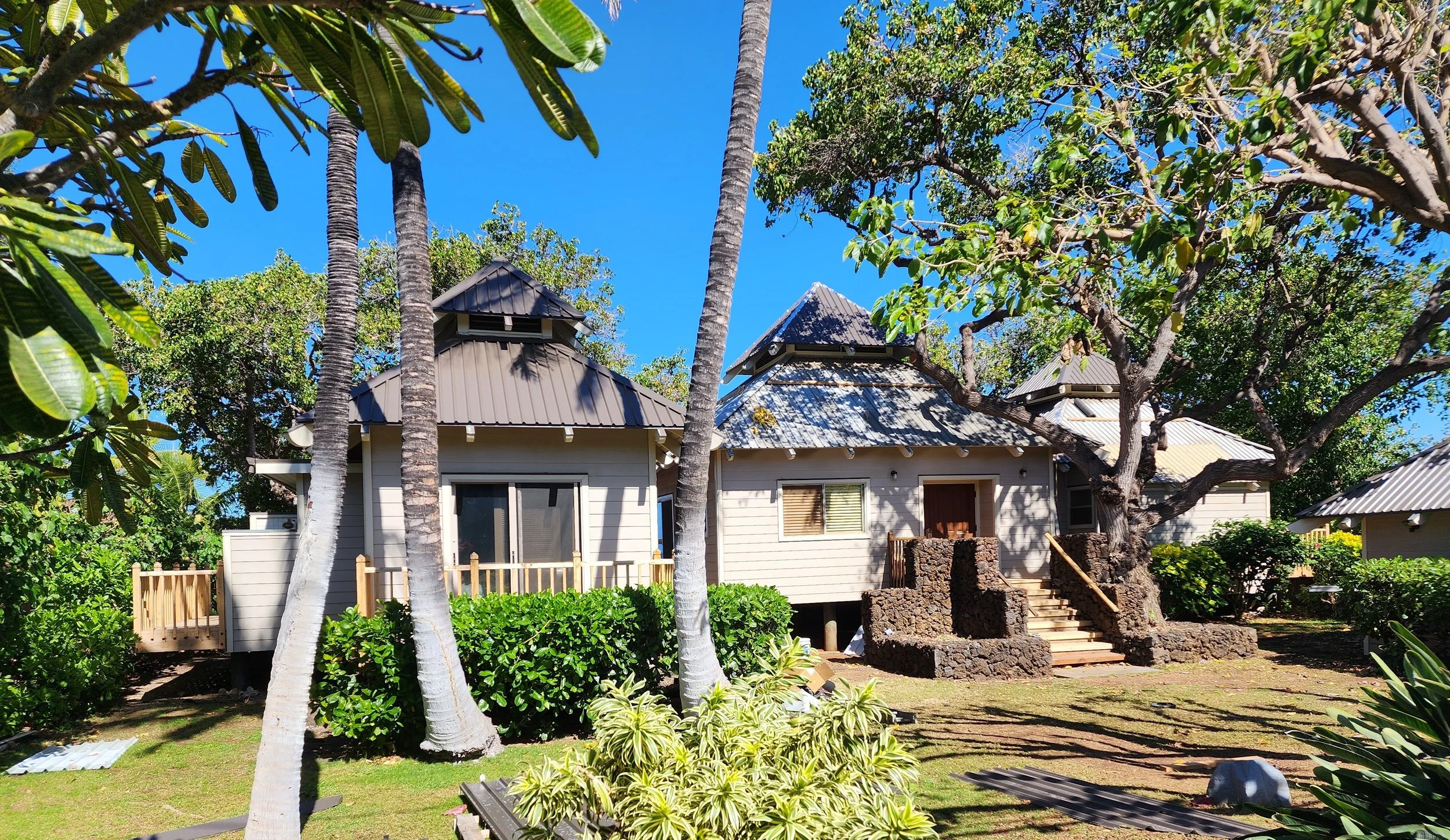 A tropical house with a tan exterior, multiple metal roofing sections, stairs leading up to the entrance, surrounded by palm trees, lush greenery, and a well-maintained lawn under a clear blue sky.