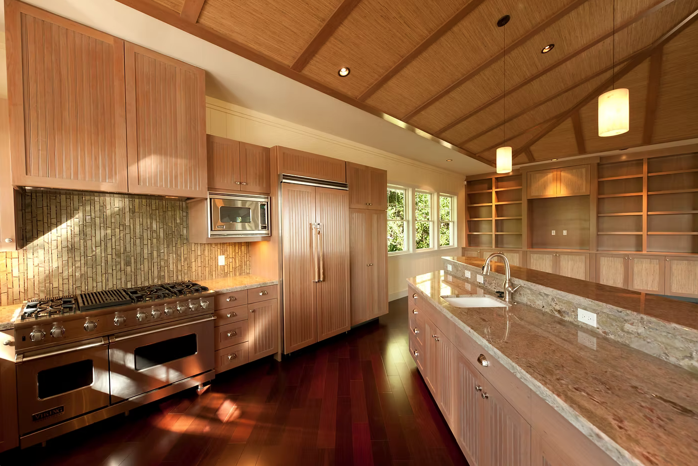 Modern kitchen with wooden cabinetry, stainless steel appliances, granite countertops, and built-in open shelves, illuminated by pendant lights and natural light through large windows.
