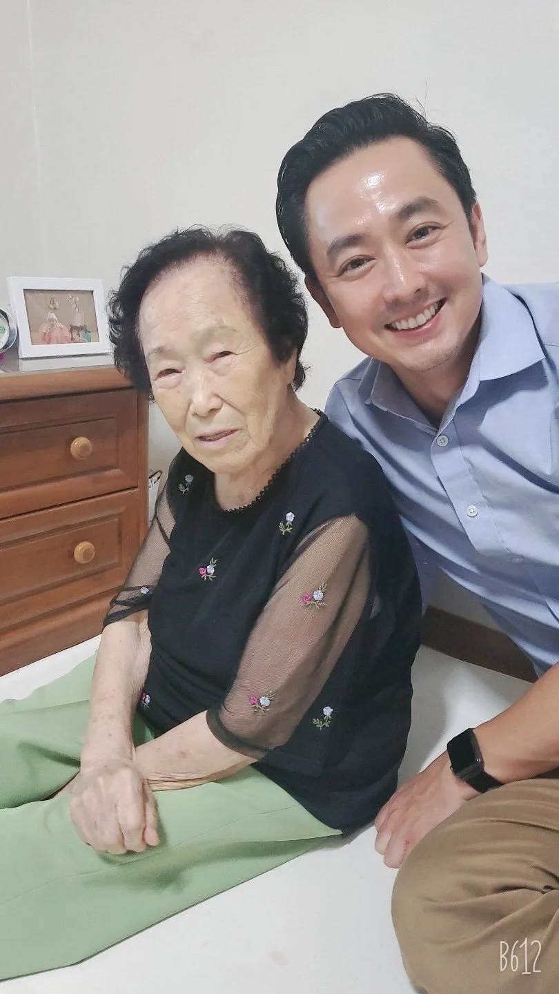 A young man with black hair and a light blue shirt smiling next to an elderly woman with black, curly hair sitting on a bed, in a room with a wooden dresser and a picture frame in the background.