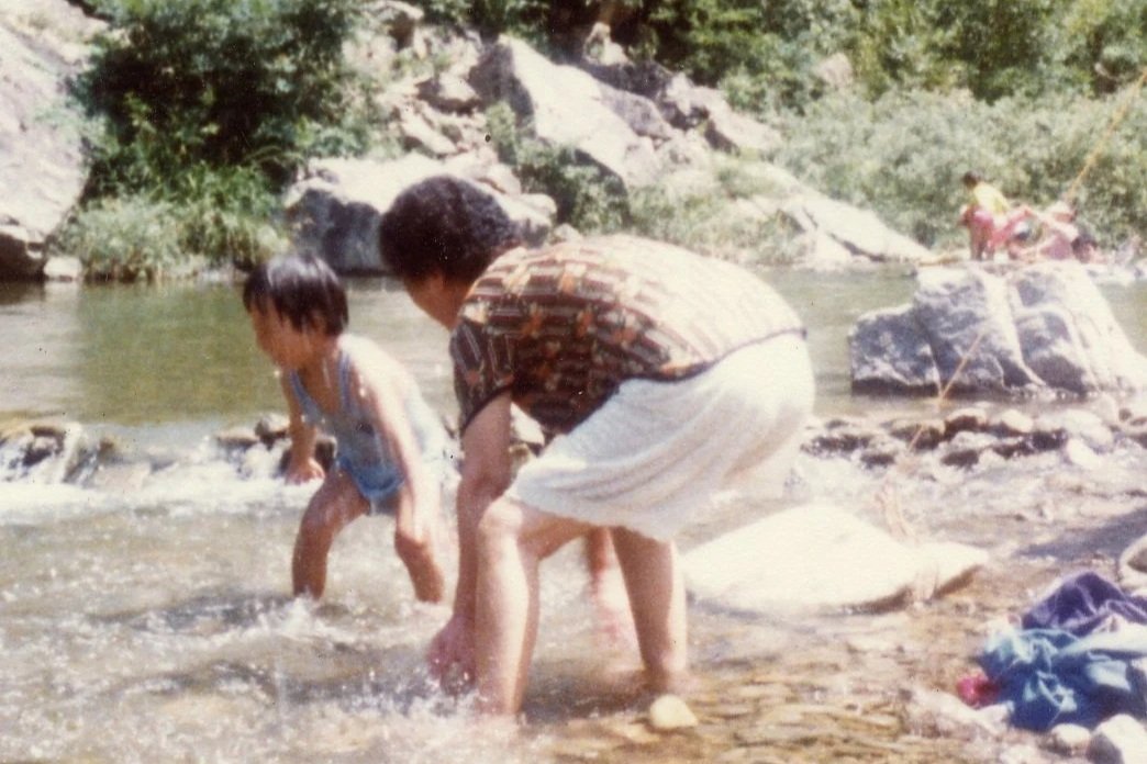A woman and a young boy playing in a shallow river with rocks, greenery, and other children in the background.