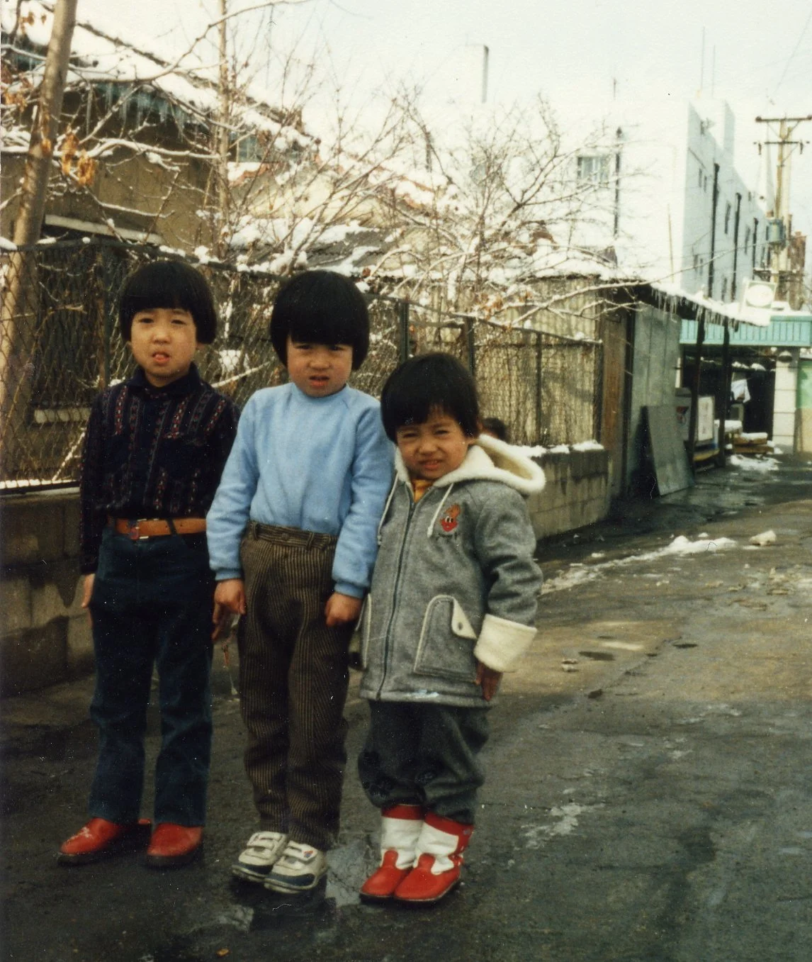 Three young children standing outdoors on a wet, snow-dusted street with houses and trees in the background.
