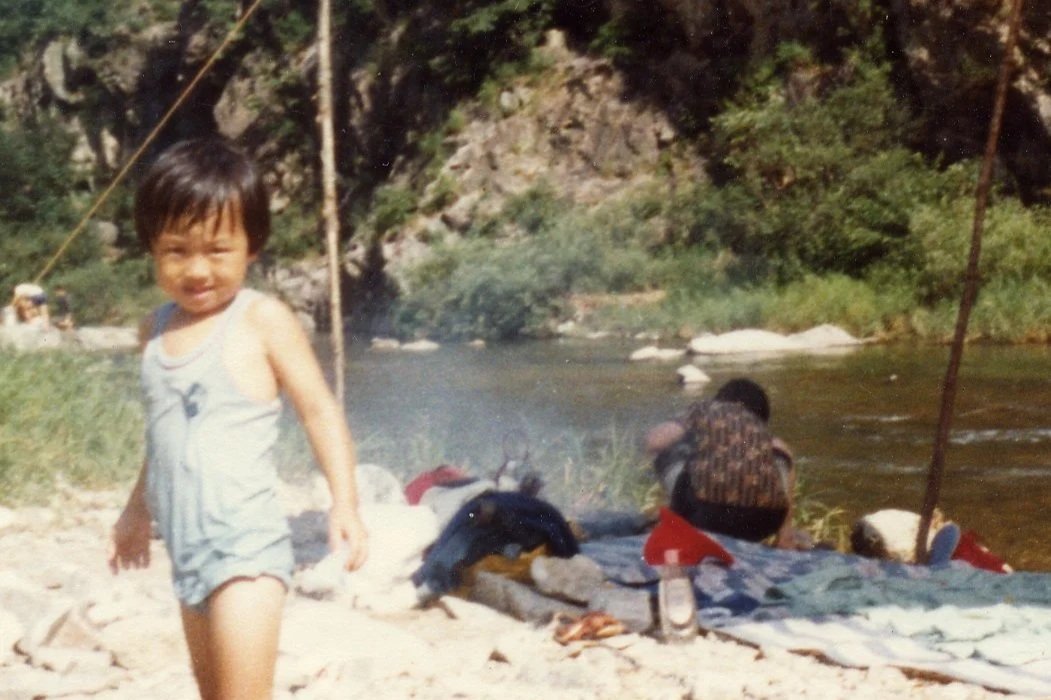 Young child smiling while walking on rocky outdoor riverside, with another person sitting by the river in the background.