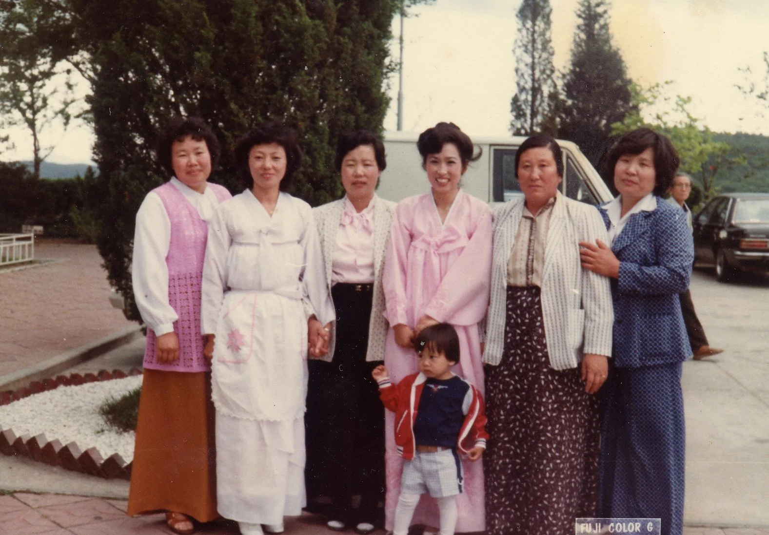 A group of seven women and one young boy posing outdoors, with the women dressed in traditional and semi-formal clothing, standing in front of trees and parked cars on a paved area.