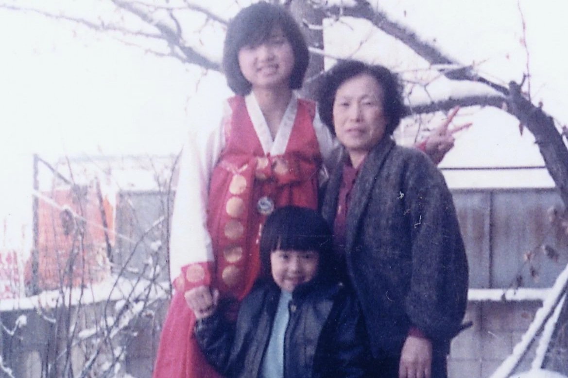 A woman and two children outdoors near a bare tree and a fence, with snow on the ground, in a winter setting.