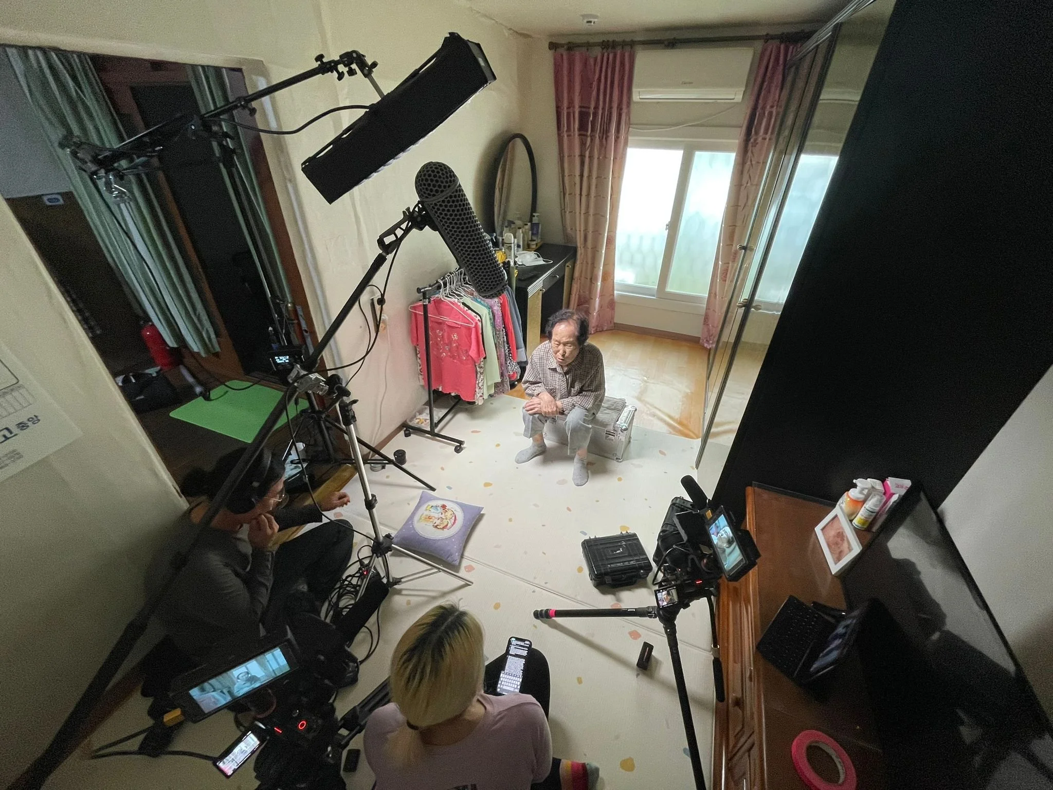 A room with a man sitting on a storage box, surrounded by filming equipment and crew, with a camera and microphone pointed at him. There are clothes on a rack, a television, and a window with pink curtains.