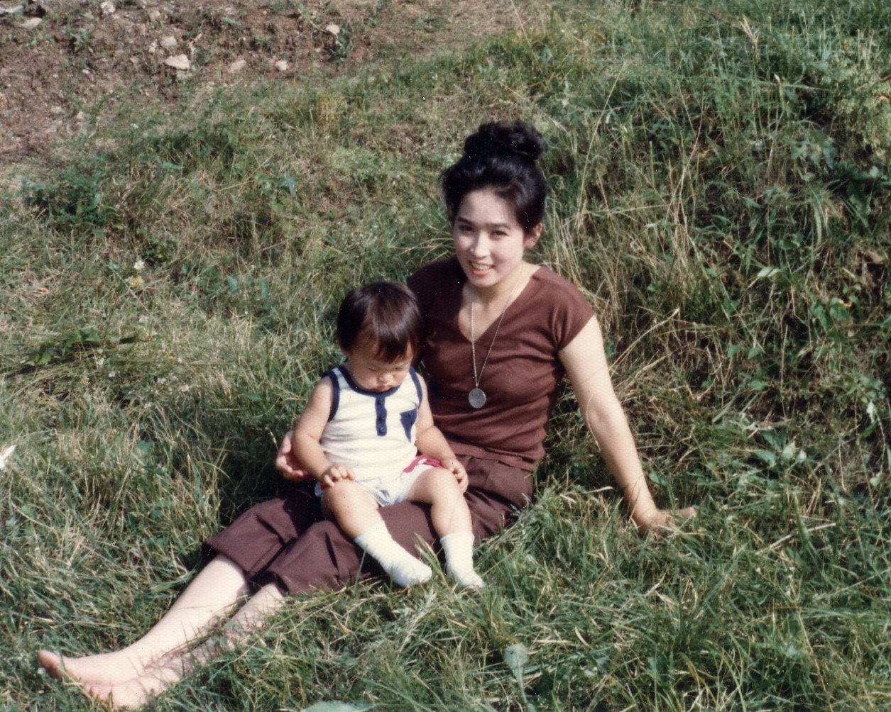 A woman with dark hair in a bun sitting on grass outdoors, holding a young child on her lap, both wearing casual clothes, surrounded by greenery.