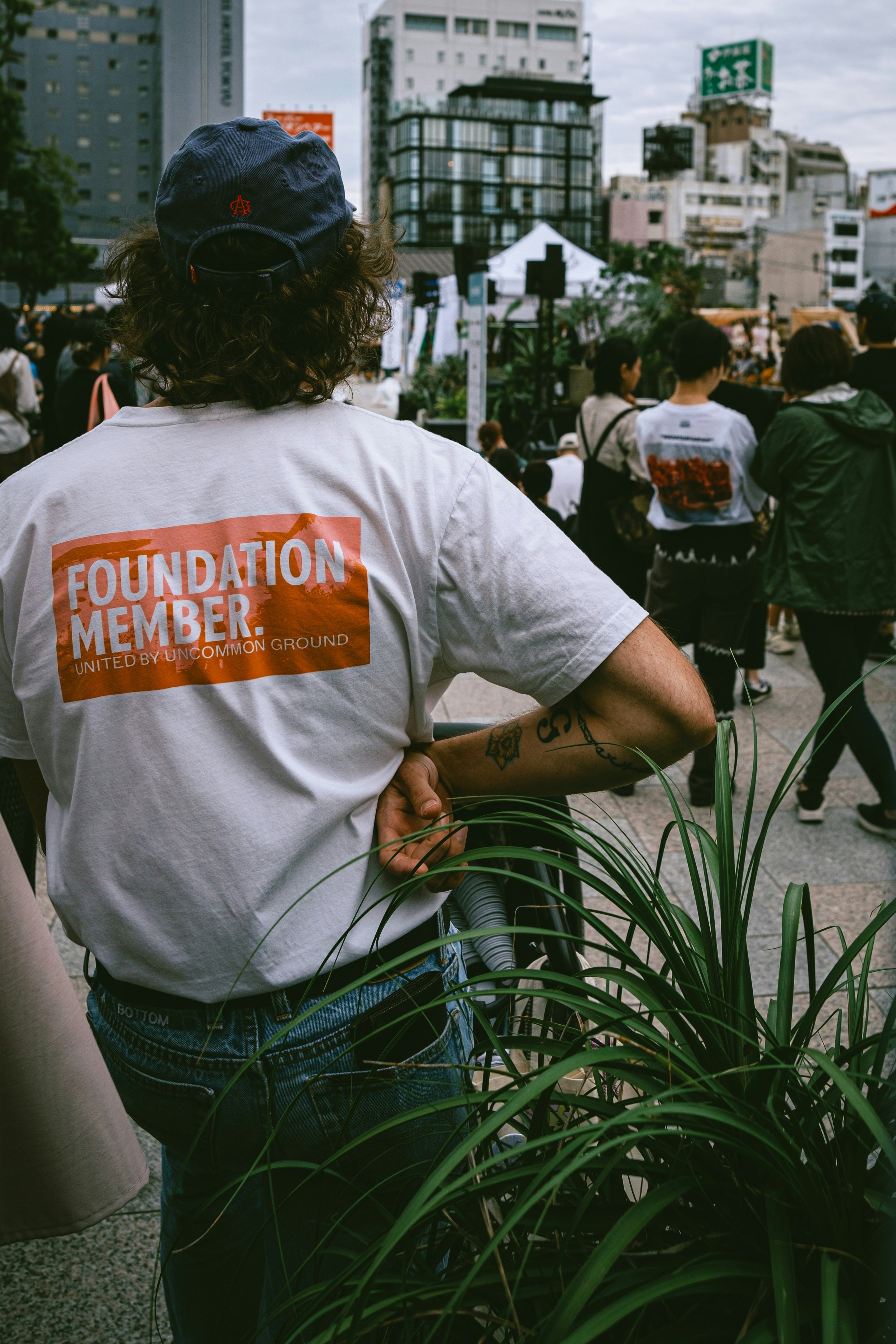 Person wearing a white t-shirt with an orange graphic reading 'Foundation Member' standing among a crowd at an outdoor event in an urban area with tall buildings.