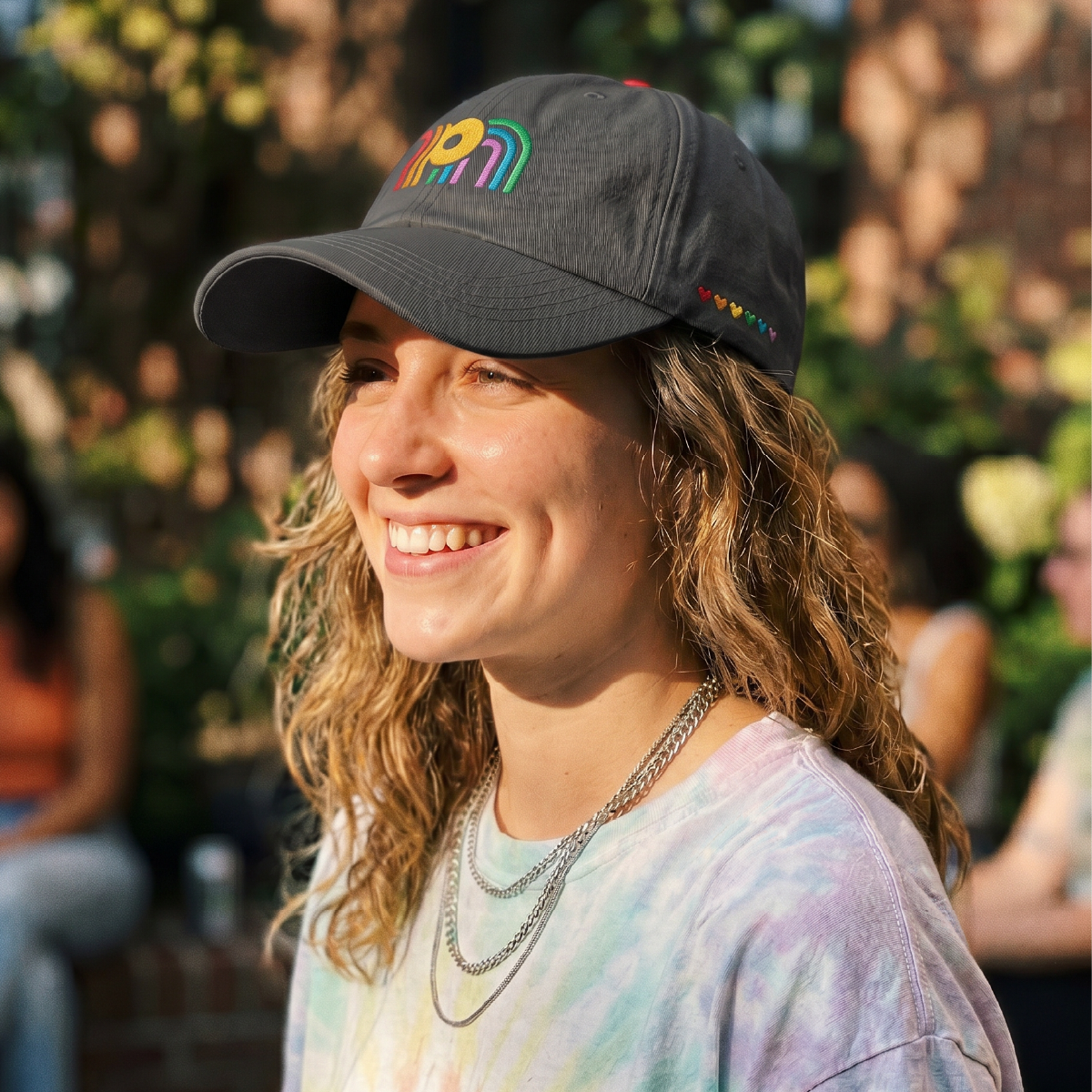 Woman with brown wavy hair smiling in sunlight, wearing a black hat with the Pride Niagara monogram logo designed by Steph Parker Designs