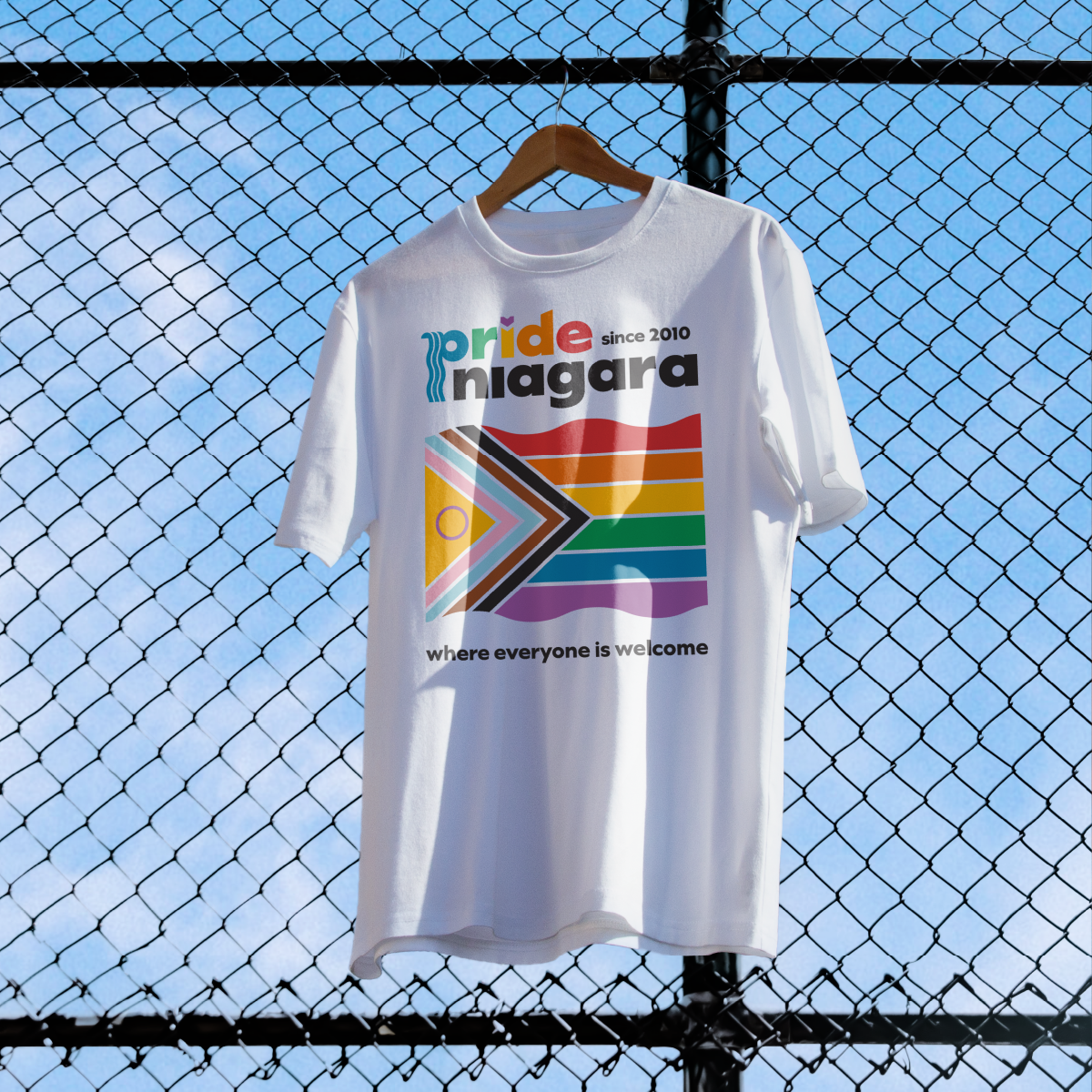 A white t-shirt with the progress pride flag, Pride Niagara logo, and the text "where everyone is welcome". Concept designed by Steph Parker Designs
