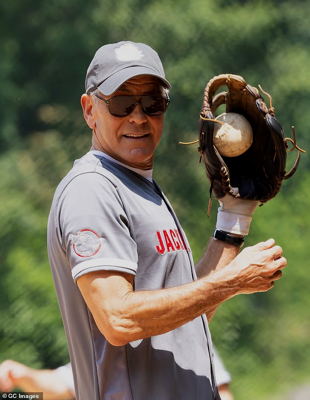 A man wearing sunglasses, a gray baseball cap, and a gray athletic shirt with red text, holding a baseball glove with a baseball in it, outdoors with a blurred green background.