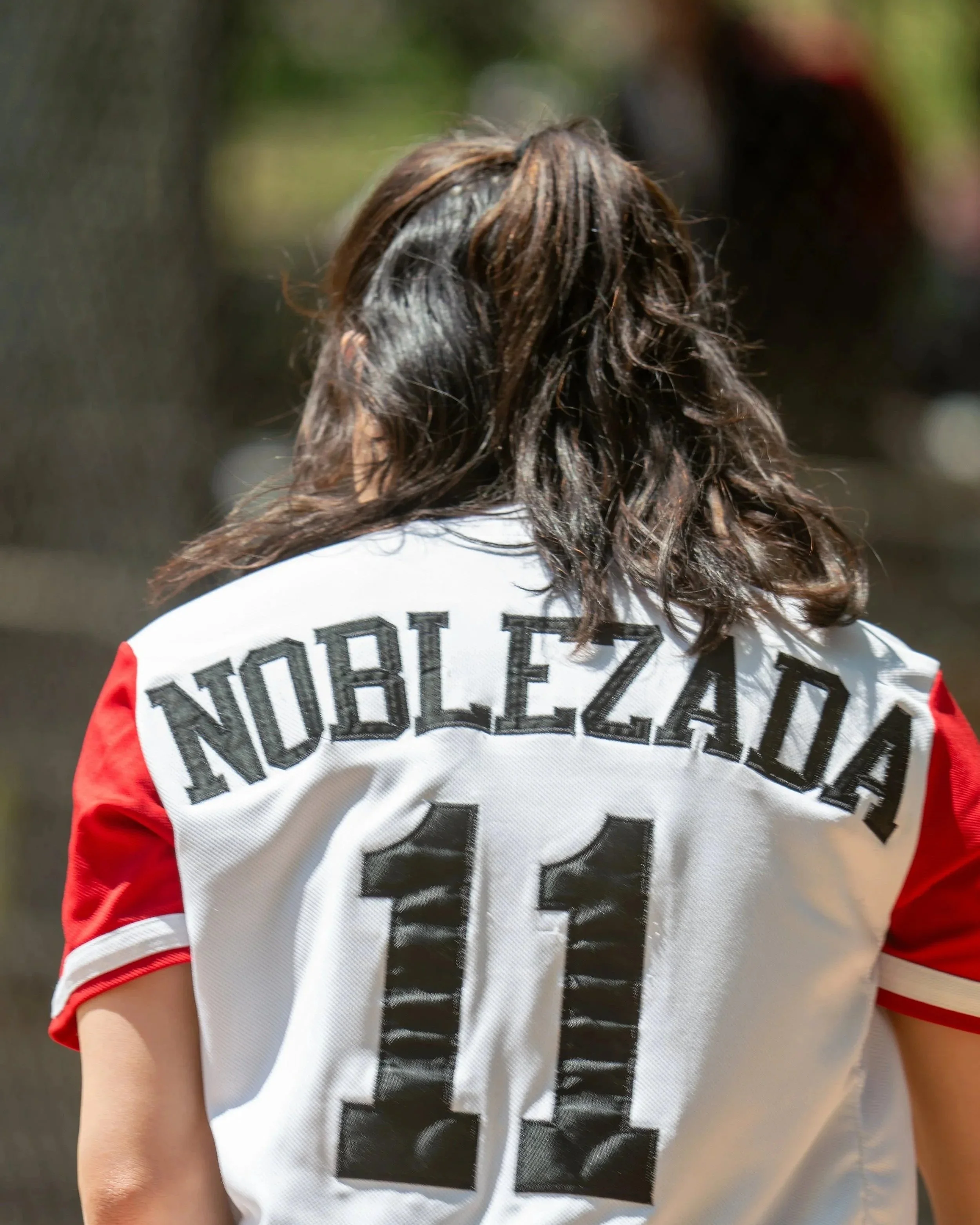 Back of a sports jersey with the name 'Nobleza' and the number 11, worn by a person with wavy, shoulder-length dark hair.