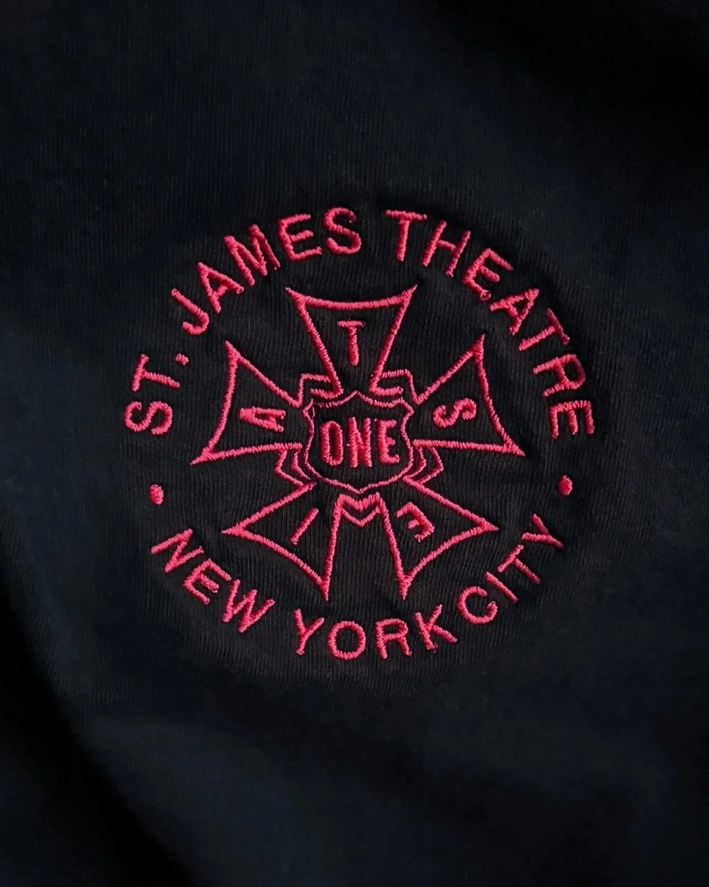 Close-up of black fabric with pink embroidered text and design. The design includes the words "ST. JAMES THEATRE," "NEW YORK CITY," and "ONE" in the center, with a star-shaped outline.