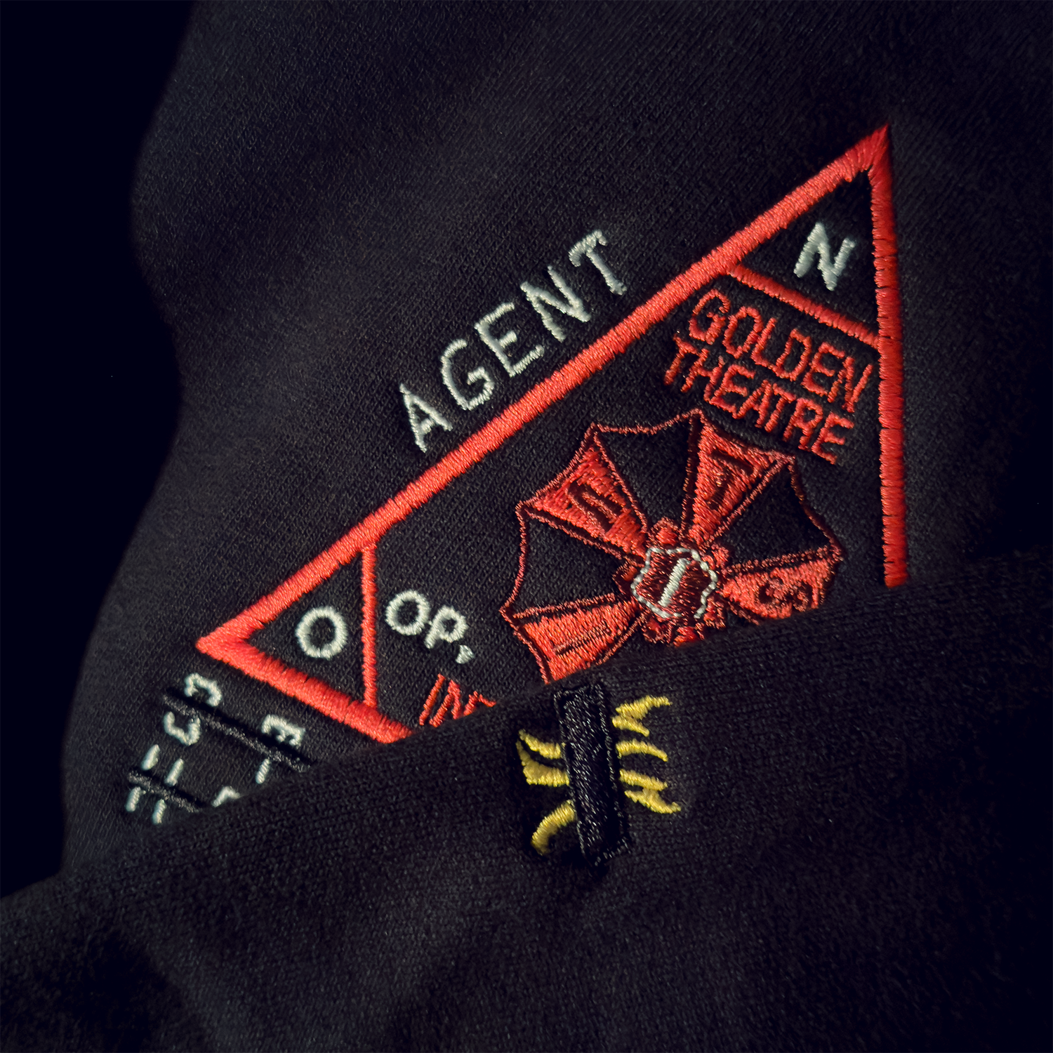 Close-up of a black embroidered patch with red, white, and yellow details, featuring a mountain logo, the words 'AGENT,' 'GOLDEN THEATRE,' and '0.,' 'O,' 'N,' and other text.