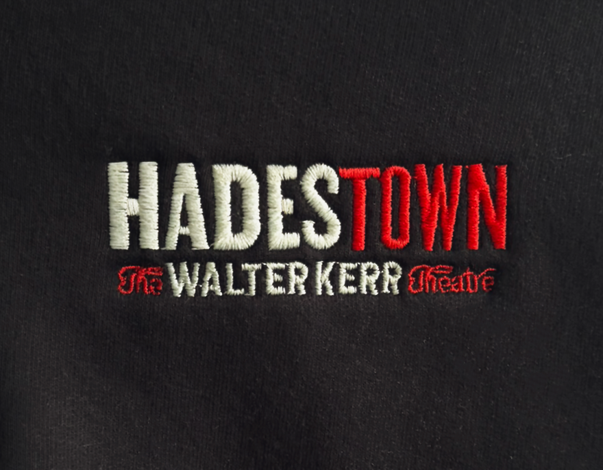 Embroidery of the words "Hades Town" in large white and red letters, with "The Walter Kerr Theatre" underneath in smaller red and white letters, on a black fabric background.