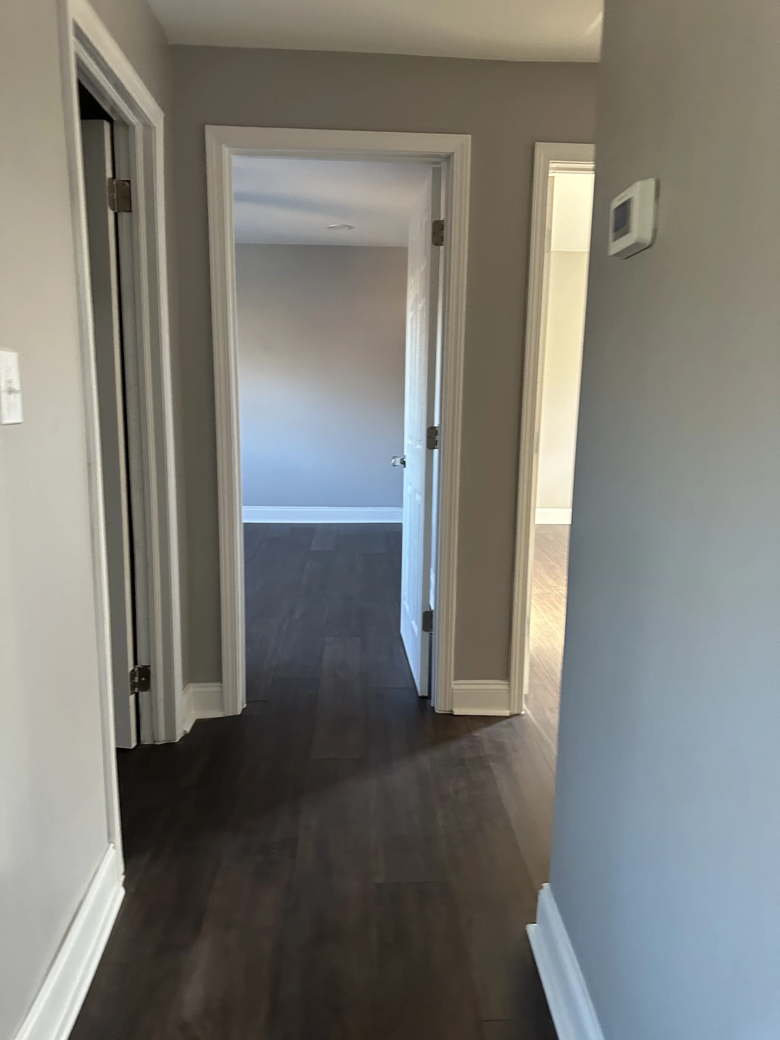 A hallway with three doorways, light gray walls, dark wooden flooring, and white trim. One door is open, leading to a room with similar flooring and light gray walls, with a closed door to the right.