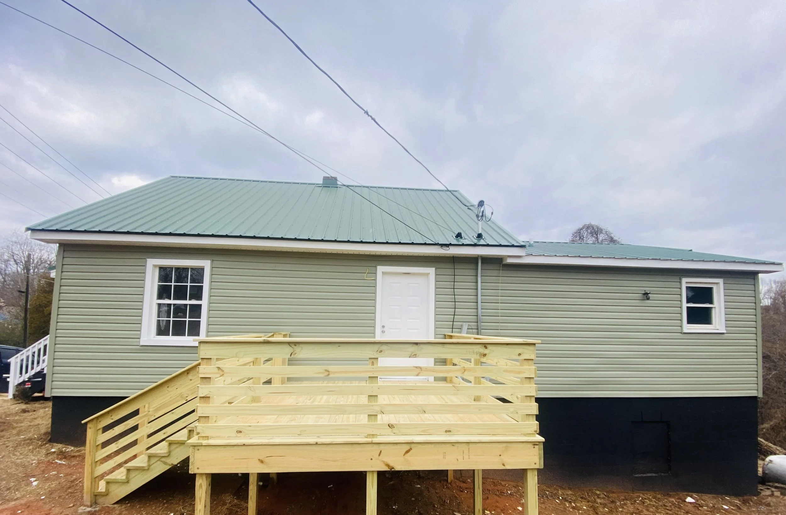 Newly constructed house with pale green siding, a metal green roof, a small wooden deck with stairs, and a white door, set against a cloudy sky.