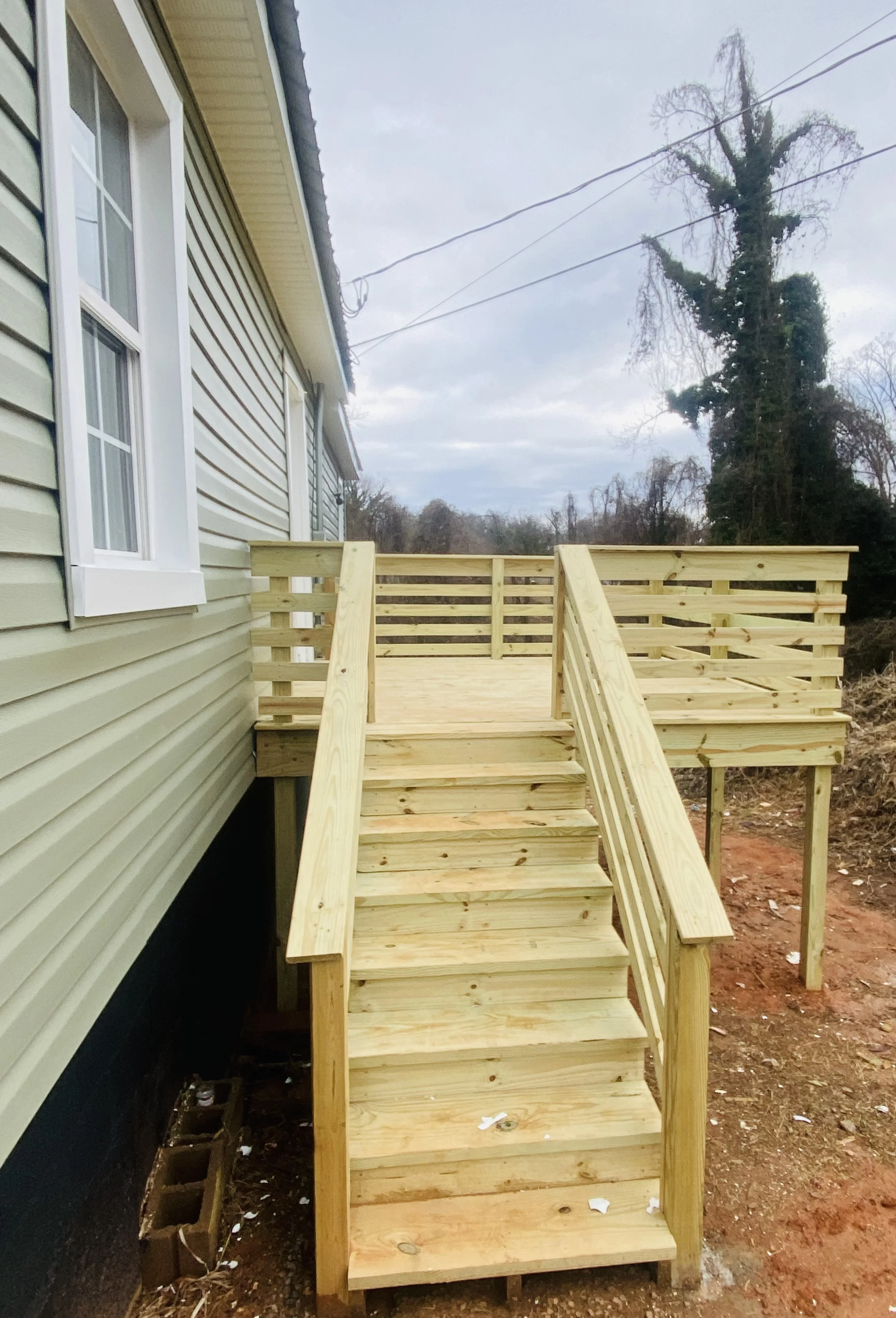 Wooden stairs and deck attached to the side of a house with beige siding, overlooking a yard with some trees and power lines in the background.