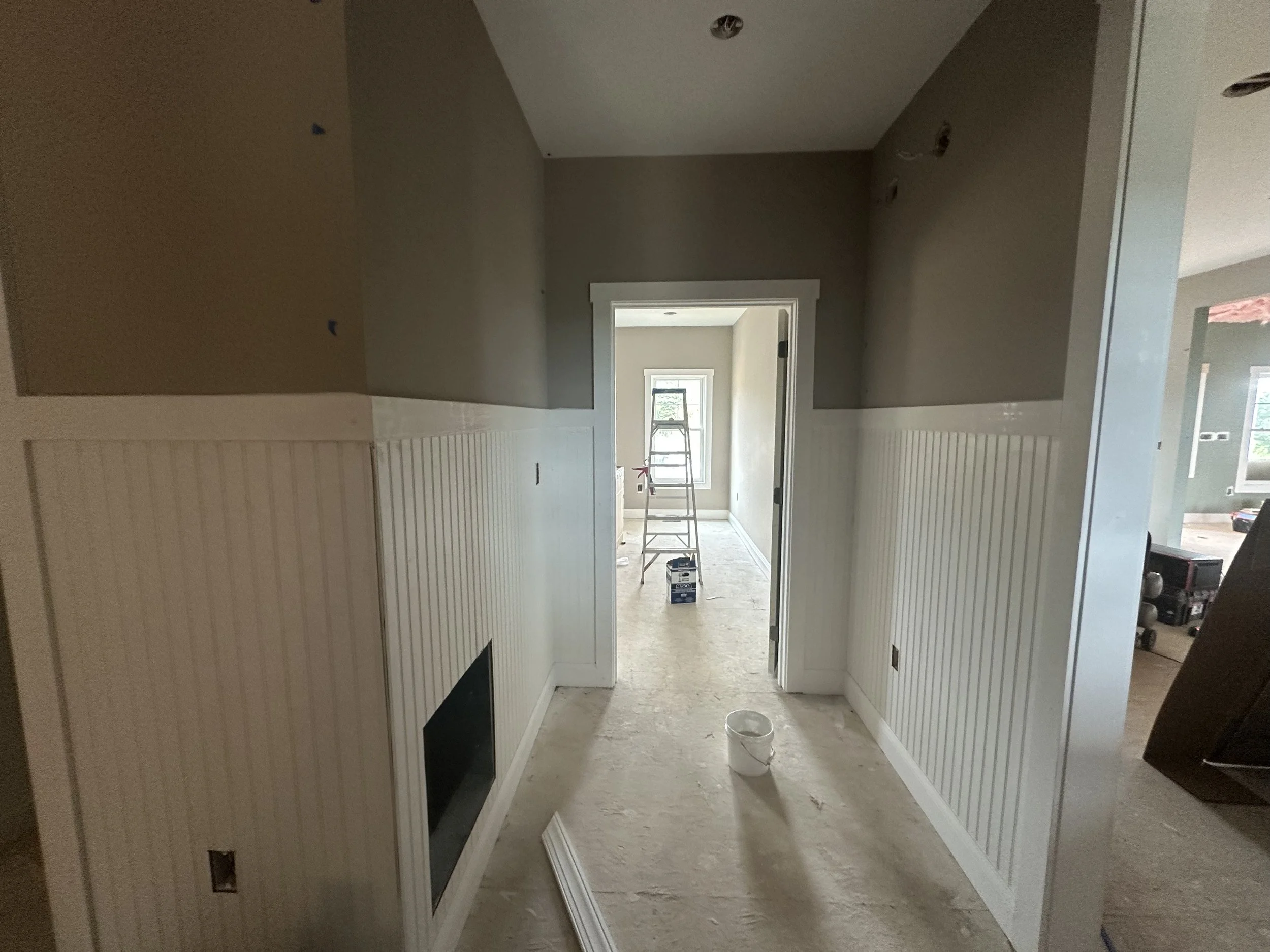 A hallway under construction with beige walls partially painted, white beadboard wainscoting, a bucket on the floor, and a ladder in the background near a window.