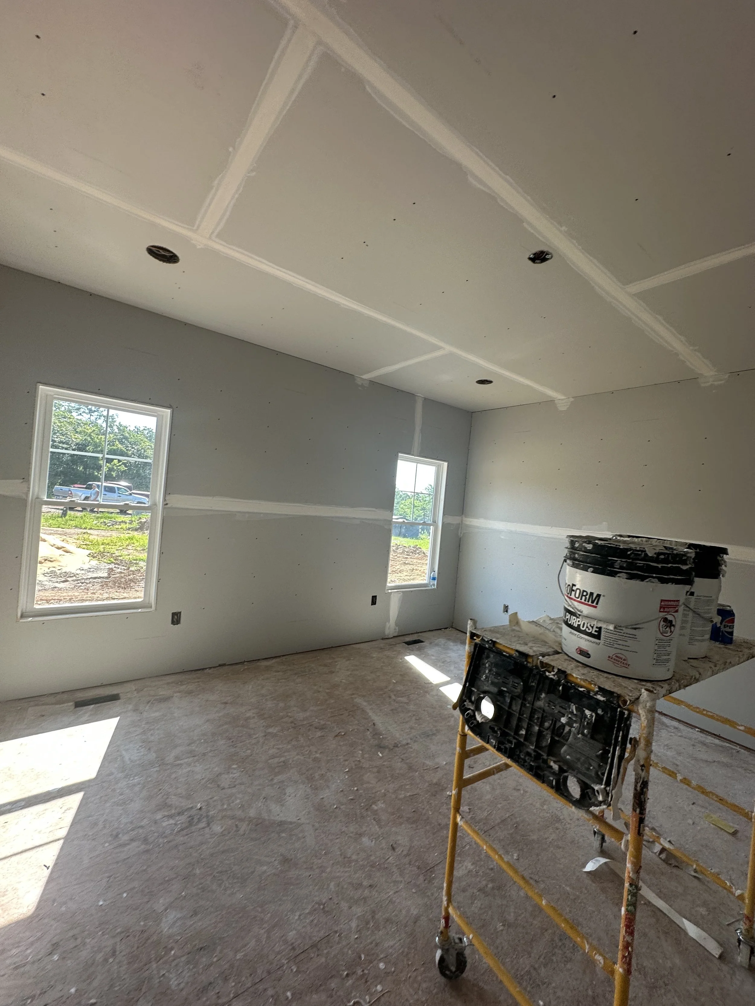 An unfinished room under construction with drywall installed on the ceiling and walls, two windows, and construction supplies including a bucket and a scaffold.