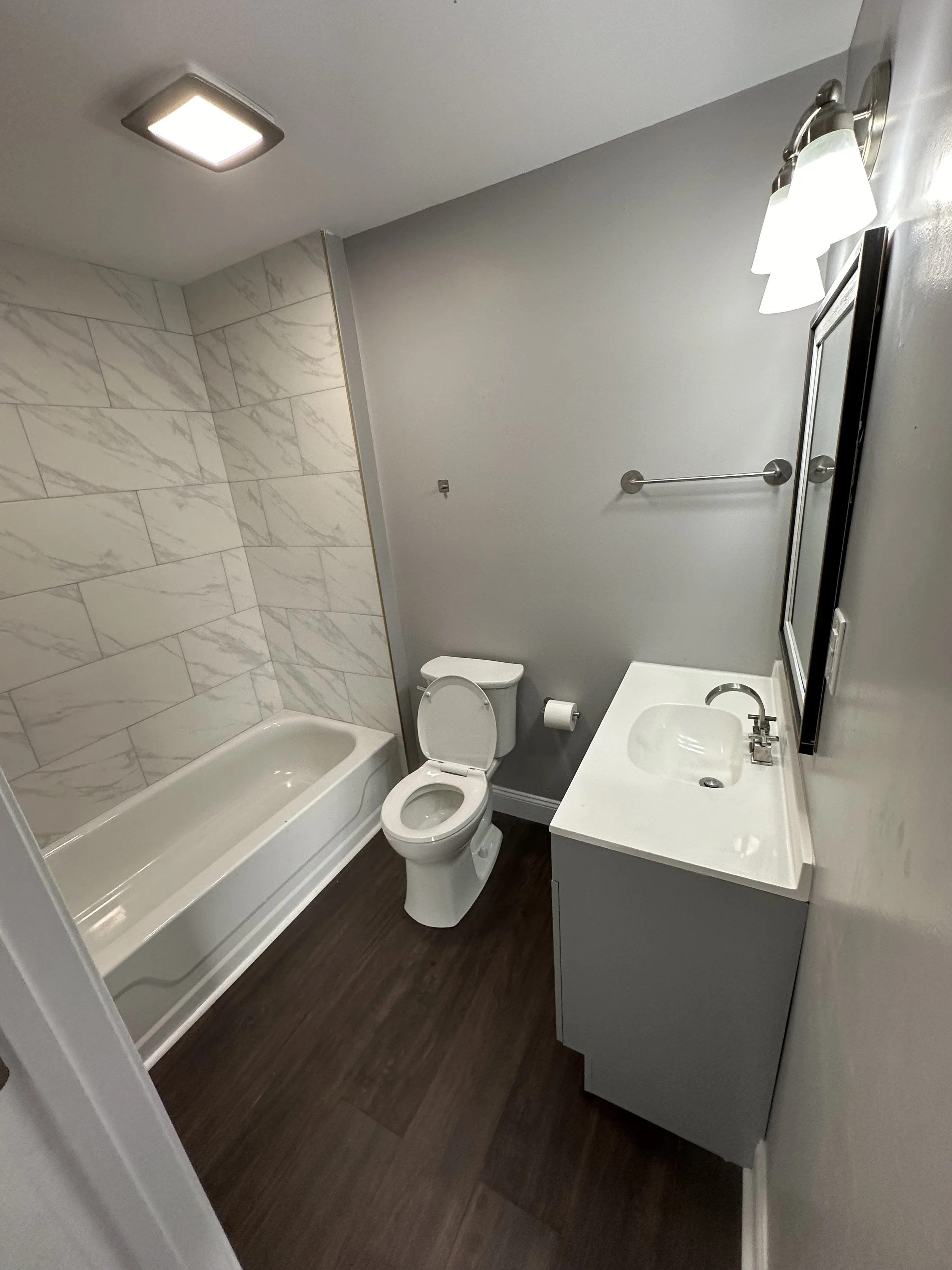 A small bathroom with a bathtub, toilet, and vanity with mirror. The bathtub has a tile wall with a marble pattern. The room has a ceiling light and wall-mounted light fixtures above the mirror.