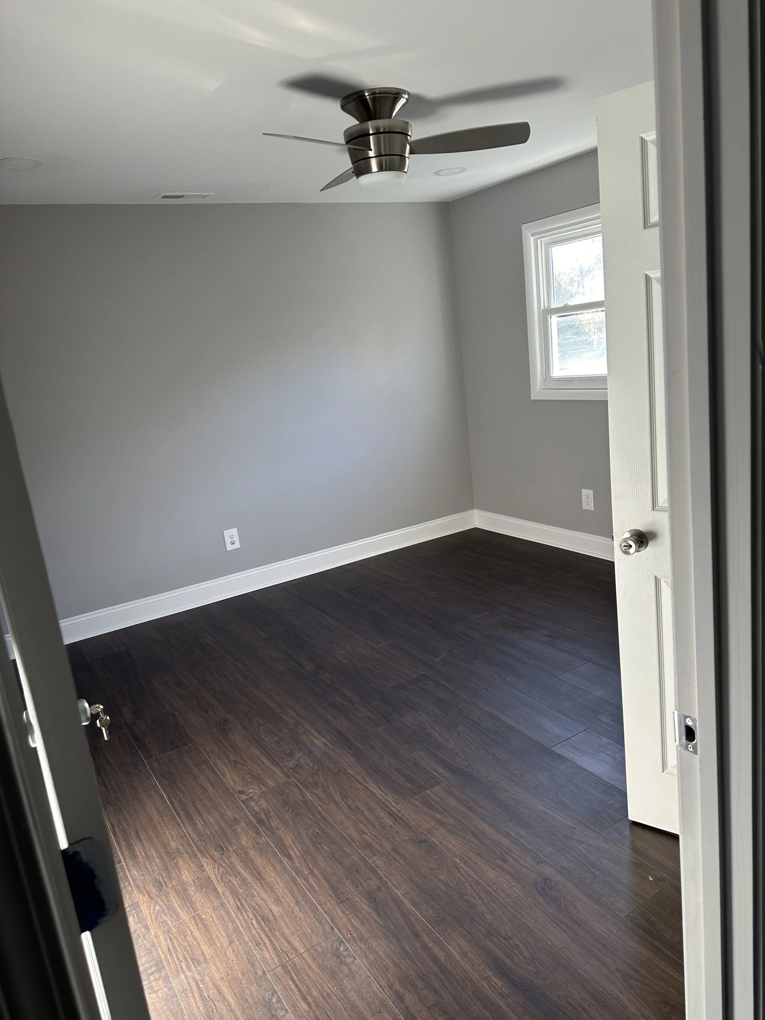Empty room with gray walls, dark wood floor, a window, a ceiling fan, and a partially open door.