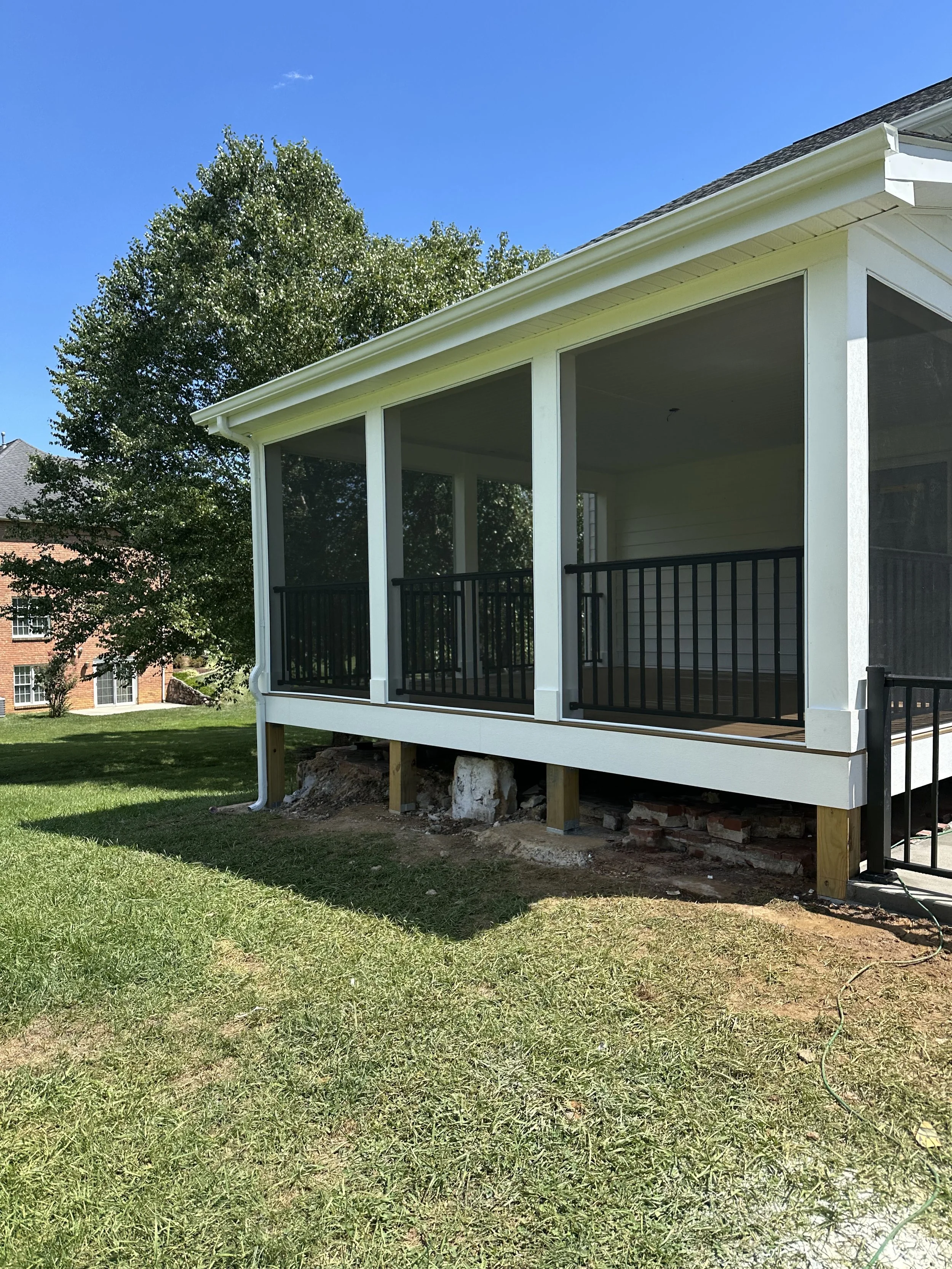 Back porch of a house with black railing, white siding, and a roof, with trees and a blue sky in the background. The porch is raised with support beams and some construction materials underneath.