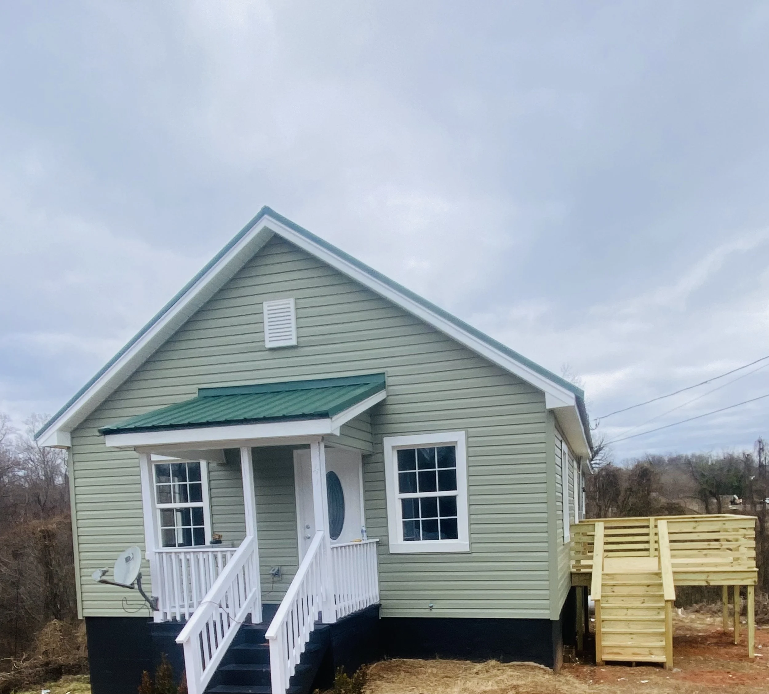 A newly built, light green house with white trim, a small front porch with white railing, and a green metal roof. There is a satellite dish attached to the porch. The house is elevated with stairs leading up to the front door, and a wooden staircase