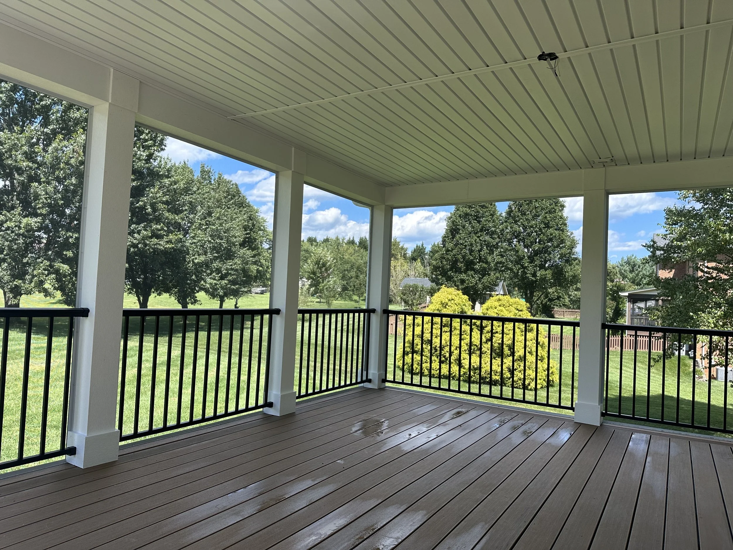 Covered porch with wooden deck, black railing, white posts, and a view of green trees and shrubs outside on a sunny day.