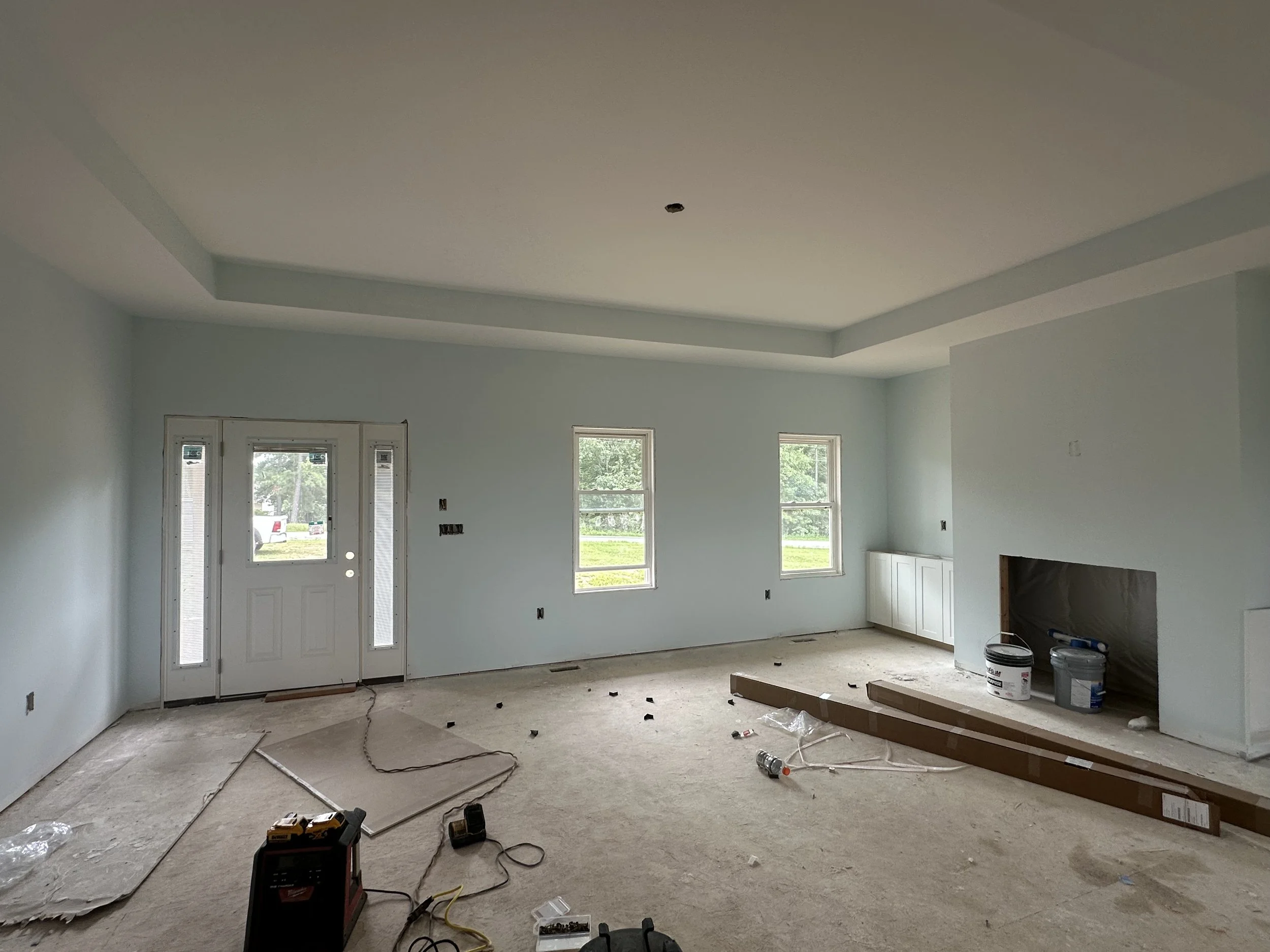 Empty living room under construction with light blue walls, three windows, and a door, with construction tools and materials scattered on the floor.