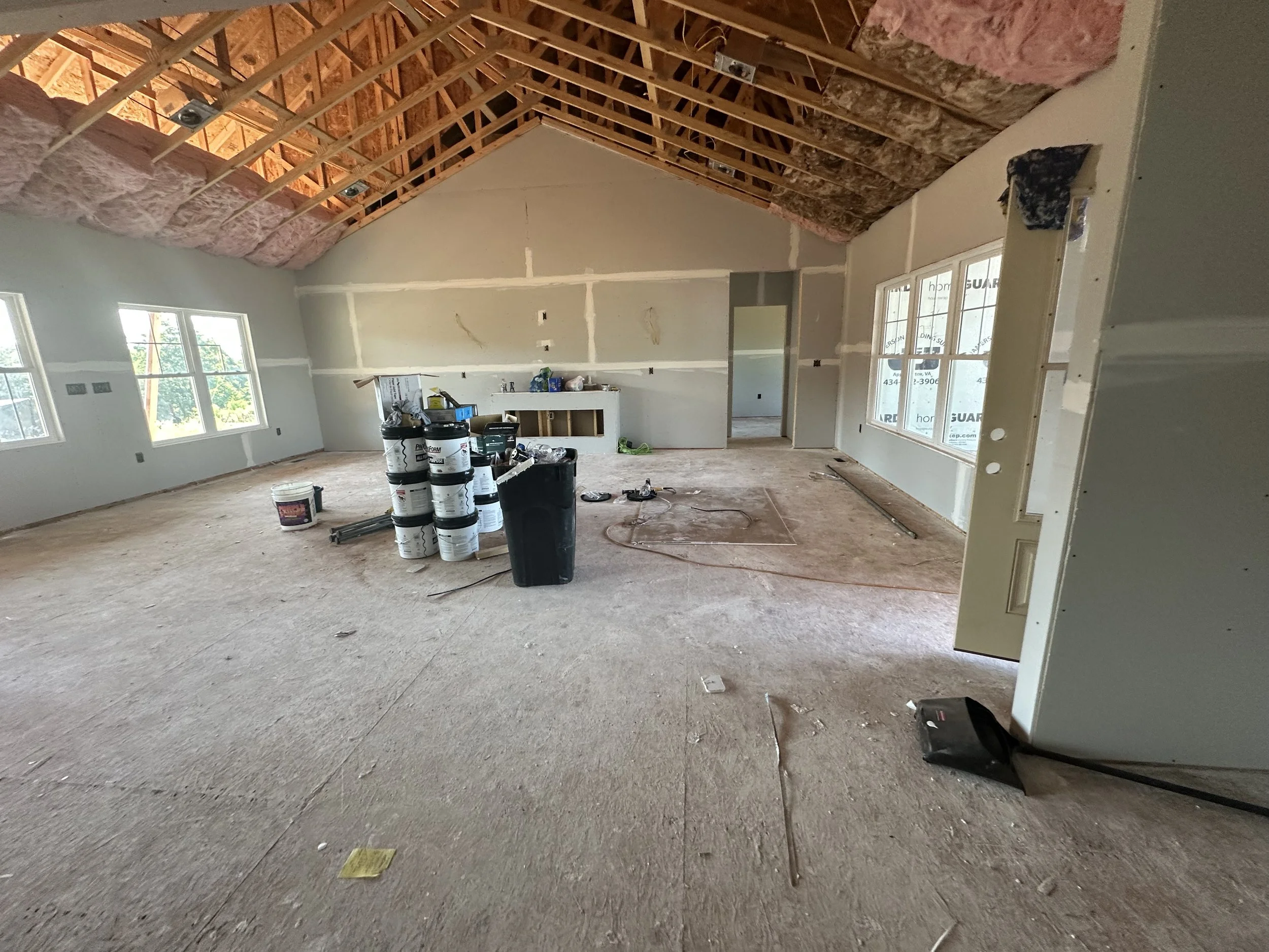 Interior of a house under construction with unfinished walls, exposed wooden roof trusses, and construction materials on the floor.