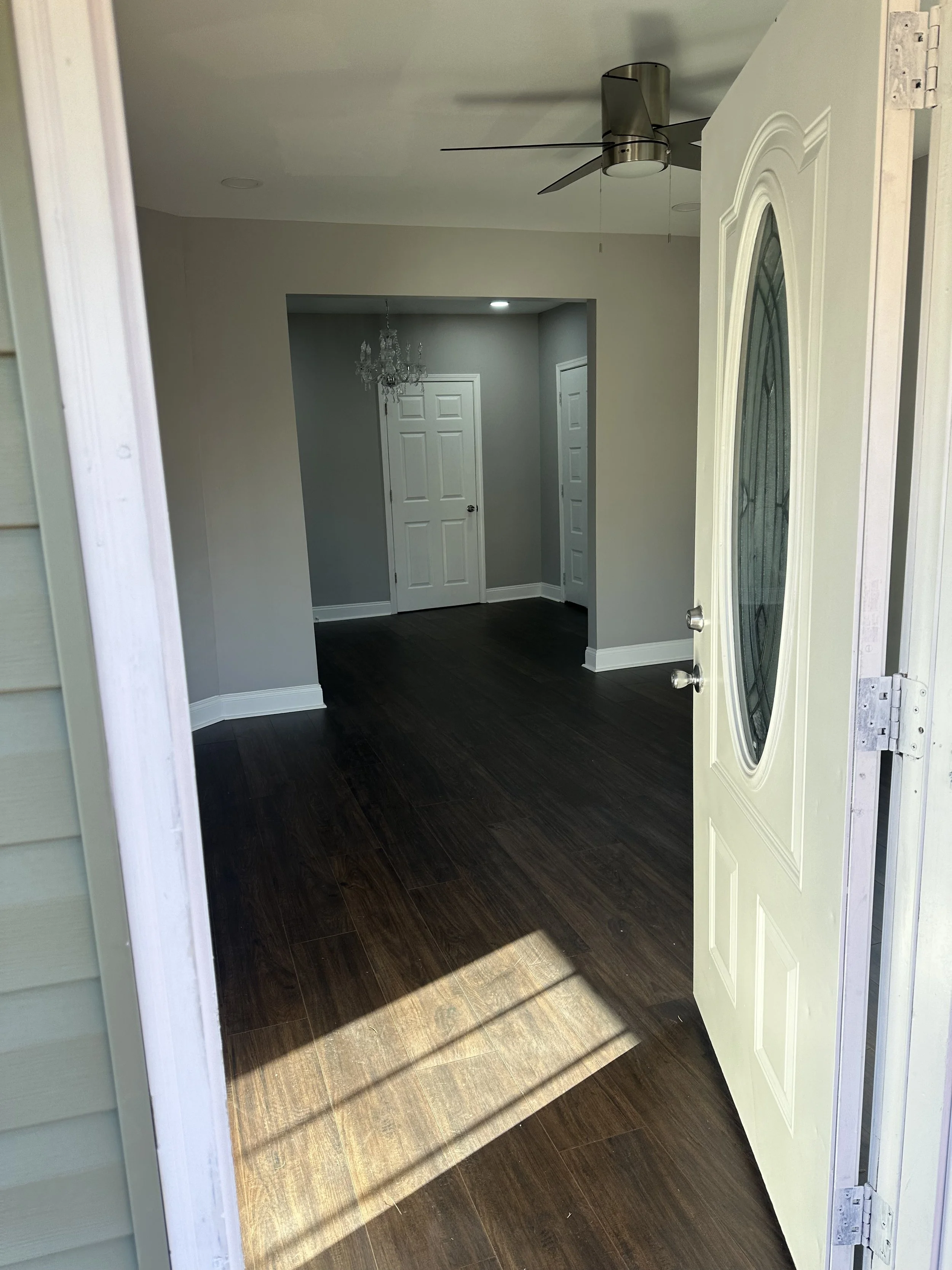 View of a house entrance with an open door showing a dark wood floor and a glimpse of a living area with a chandelier ceiling light, a ceiling fan, and gray walls.