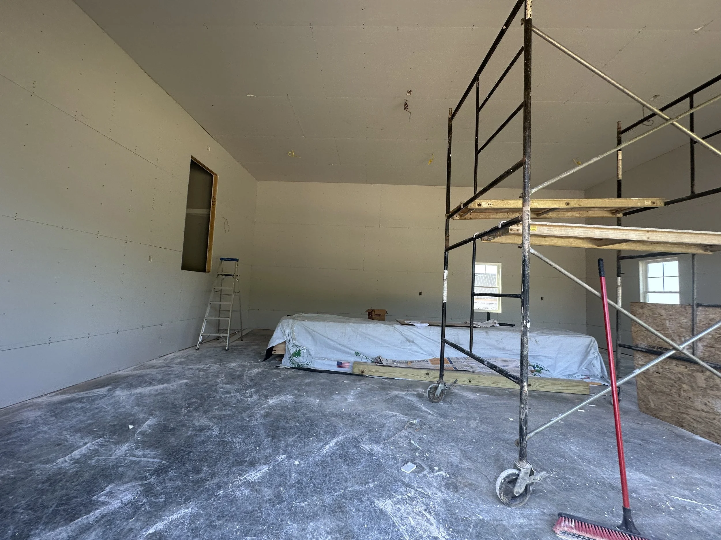 Interior of a room under construction with unfinished drywall walls, a ladder, a scaffold, a covered work surface, and construction tools on the floor.