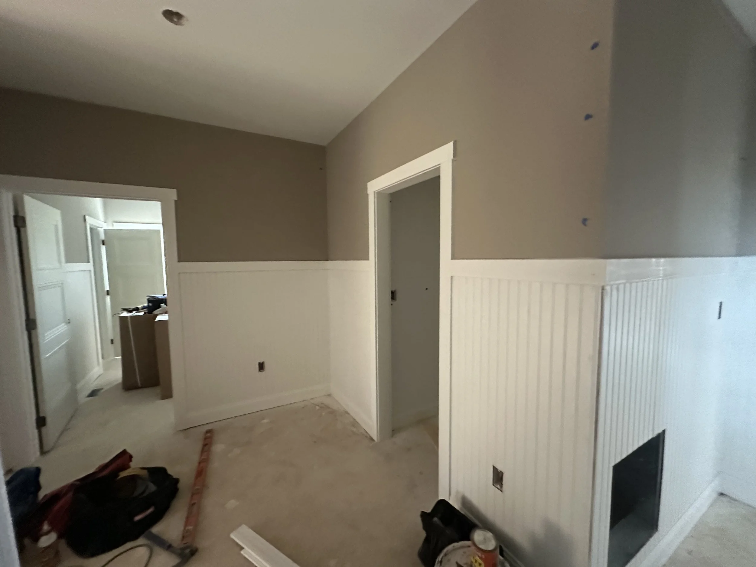 Interior of a room under construction with beige walls, white wainscoting, and doorways, construction tools and supplies on the floor.