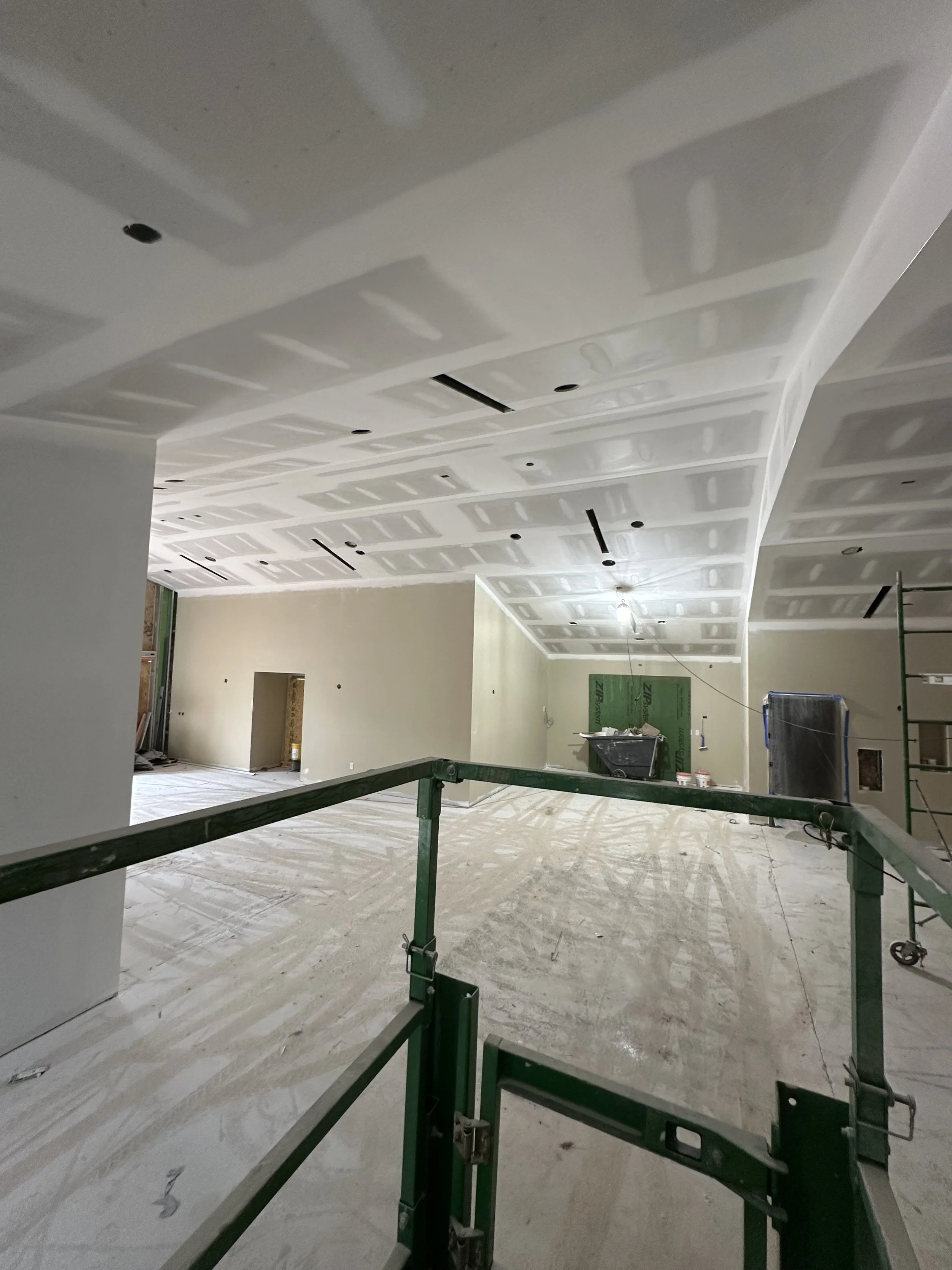 Interior of a building under construction with drywall in the process of being installed on the ceiling and walls, scaffolding in the foreground, and construction materials scattered on the floor.
