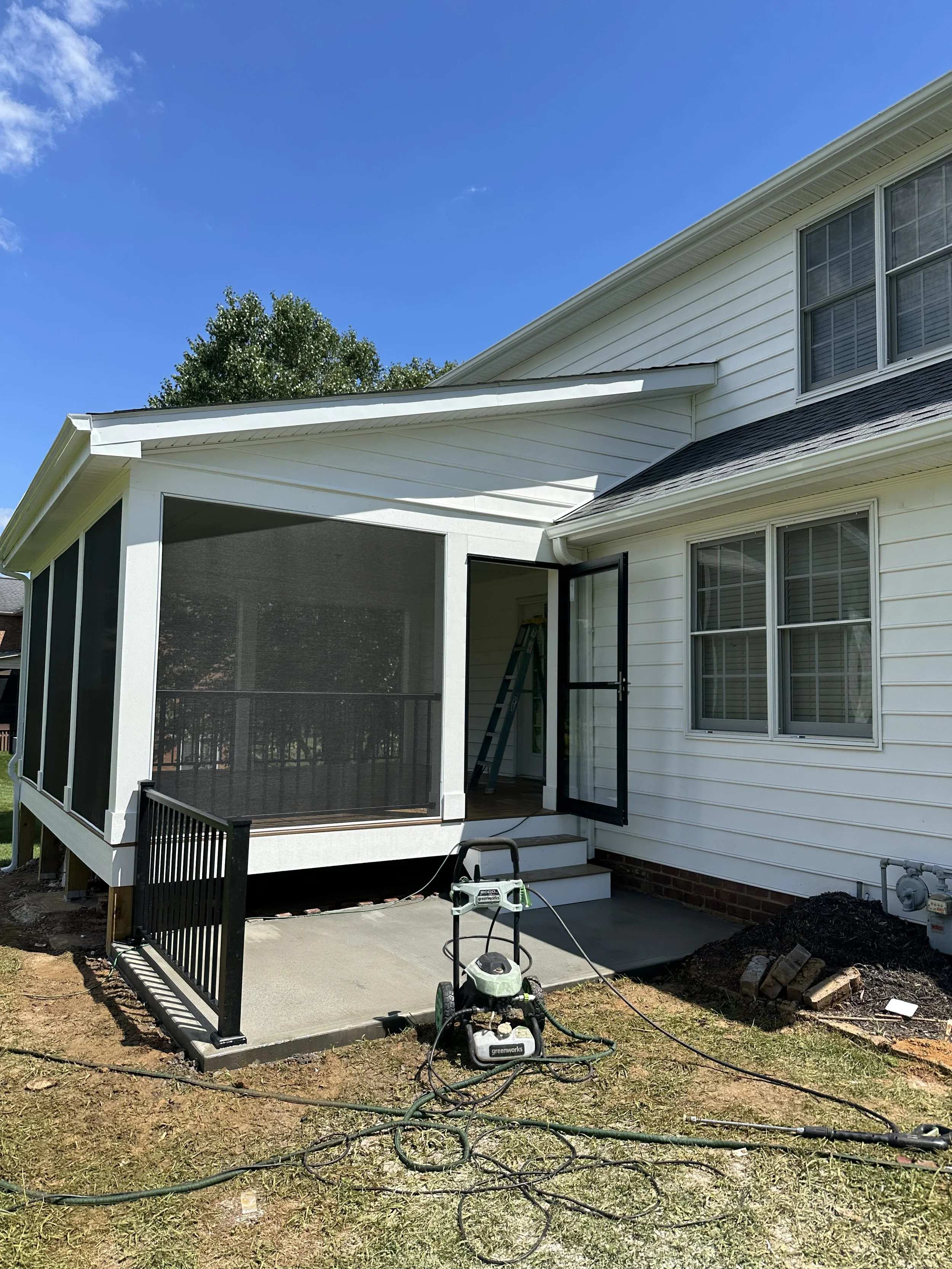 Newly constructed screened-in porch attached to a white house with steps leading up to a door, construction equipment and tools in front, and a bright blue sky overhead.