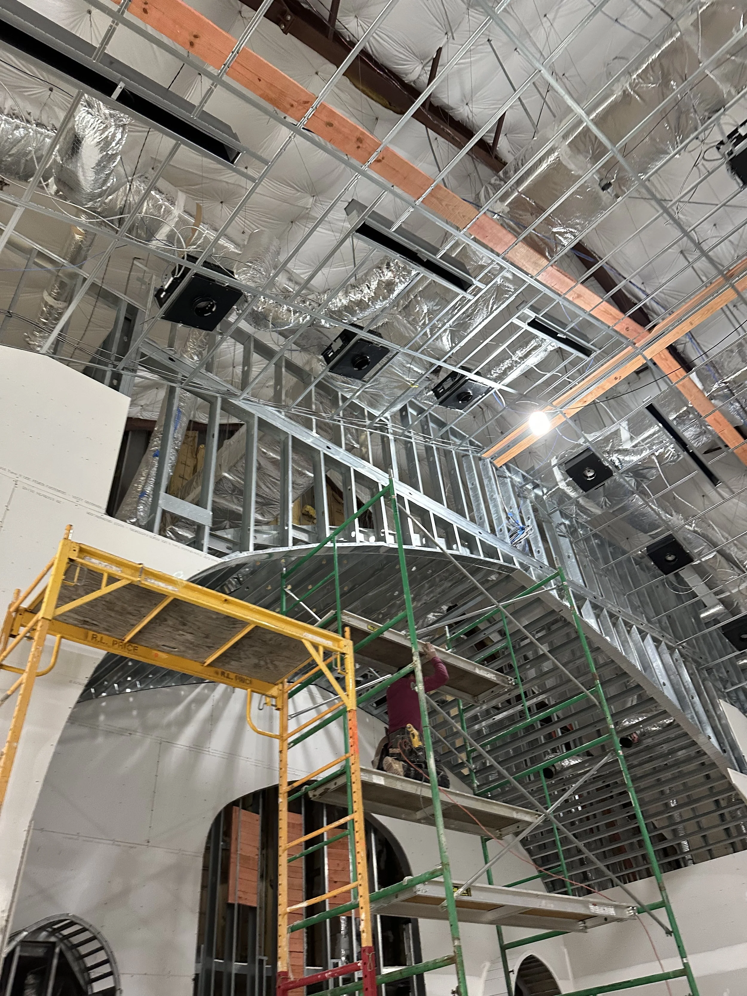 Construction worker building a curved metal staircase inside a building with exposed ceiling ducts and framing.